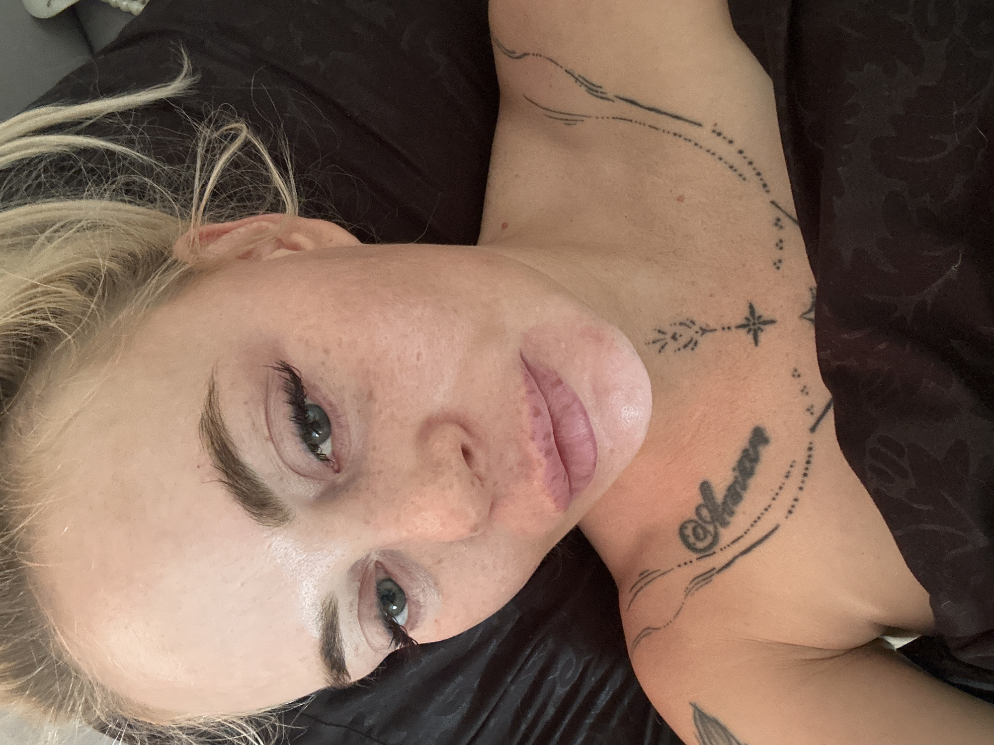 A close-up overhead shot shows a fair-skinned woman with blonde hair and green eyes, lying down. Her eyes are open, and she is looking upwards at the camera. Her makeup includes false eyelashes and lip filler. She has freckles and some redness on her face. A black tattoo is visible on her upper chest and shoulder. The background is a dark, textured fabric, possibly a pillow or blanket. The lighting suggests an indoor environment. The image was taken at an unknown location in Unknown, Unknown.