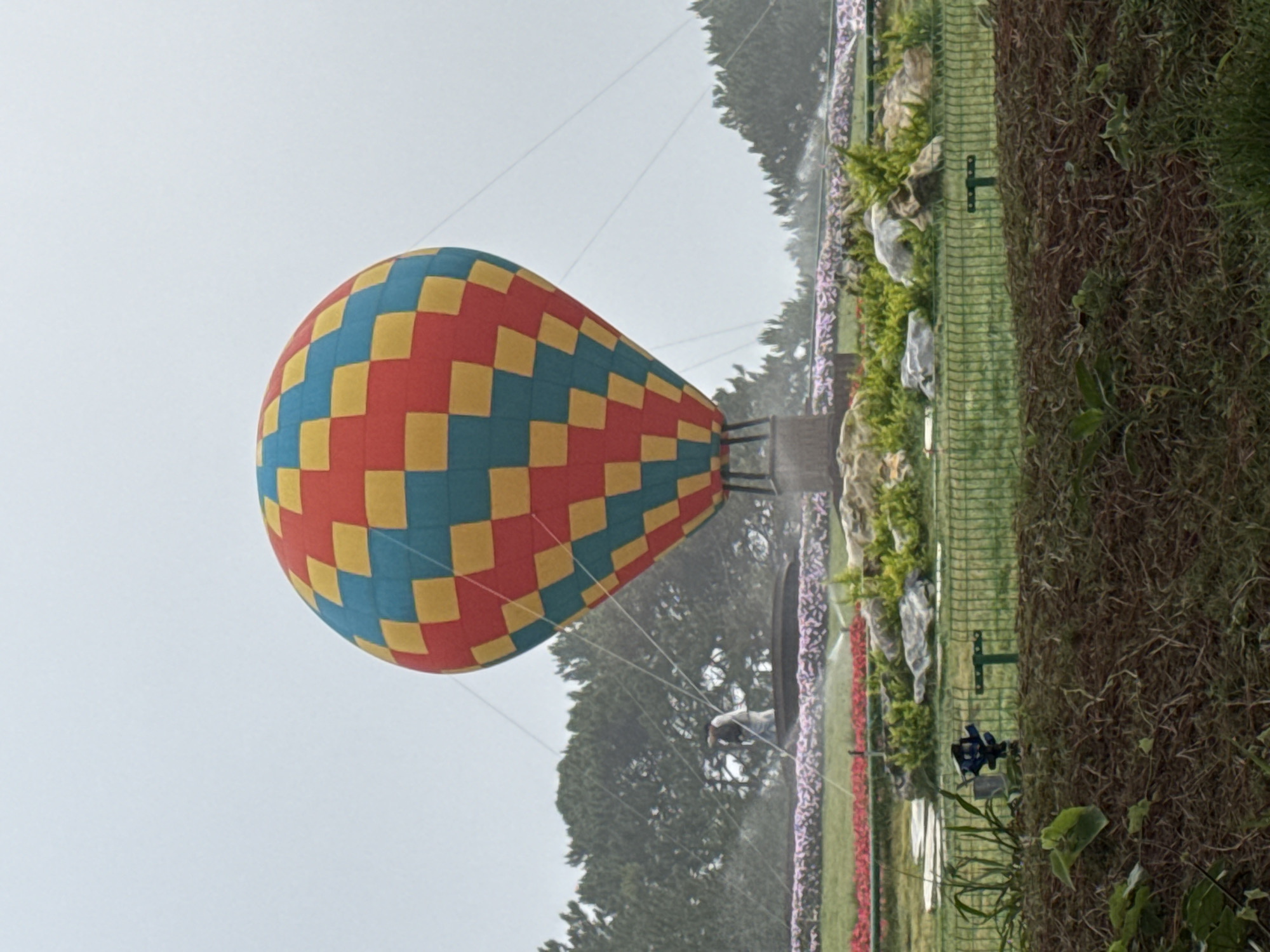 A colorful hot air balloon is displayed on the edge of an artificial waterfall at a fair in the city of Da Lat, Vietnam. The balloon is composed of red, yellow, and blue squares arranged in a checkered pattern. The background includes tall trees and a cloudy sky. A display of pink, red, and purple flowers lines a green fence that is backed by brown foliage.