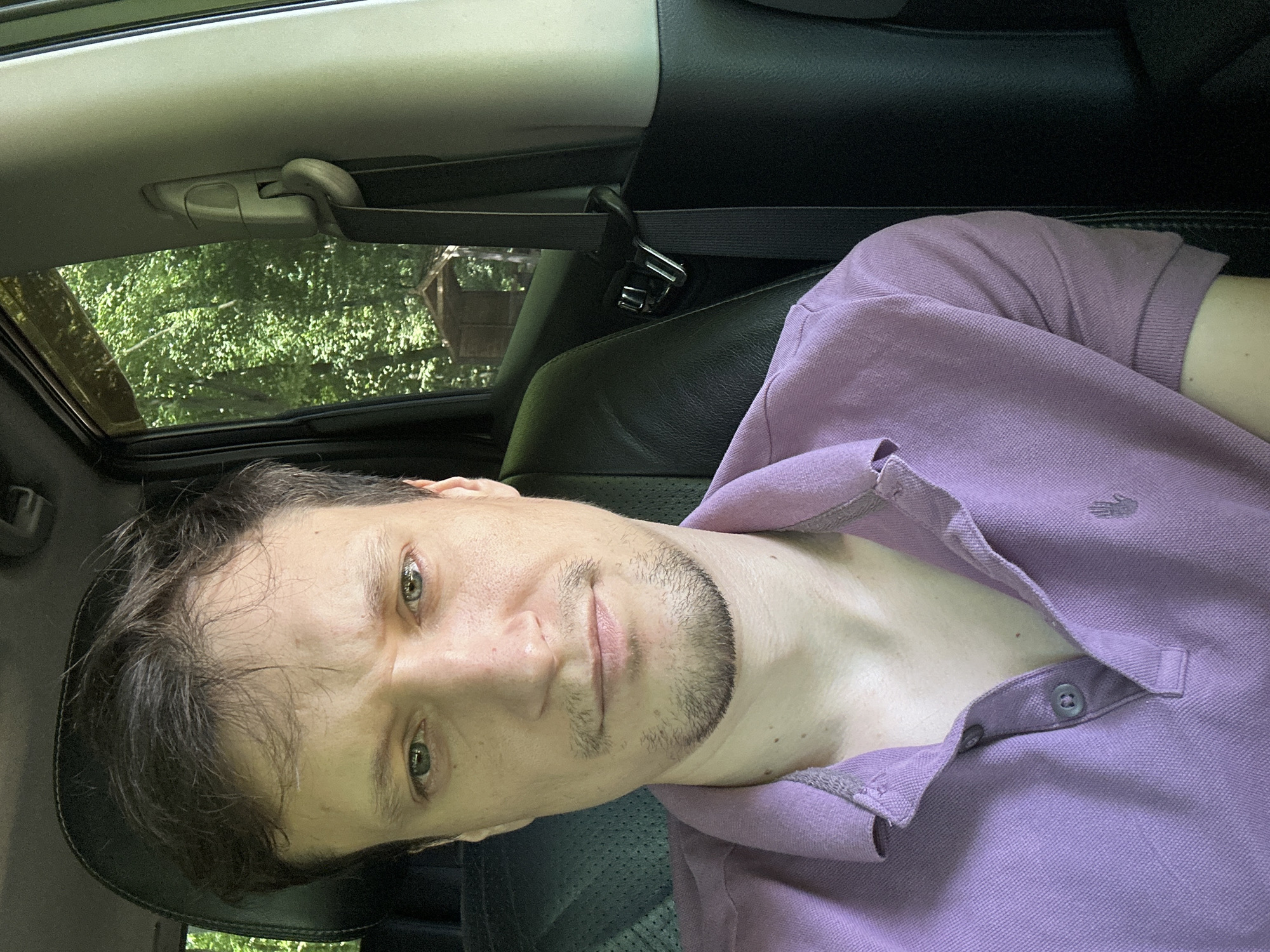 A man is sitting in the passenger seat of a car. He is looking at the camera. He has short brown hair and a light beard. He is wearing a purple polo shirt. The car is parked on a road with trees and a small building in the background. It is daytime and the sun is shining.