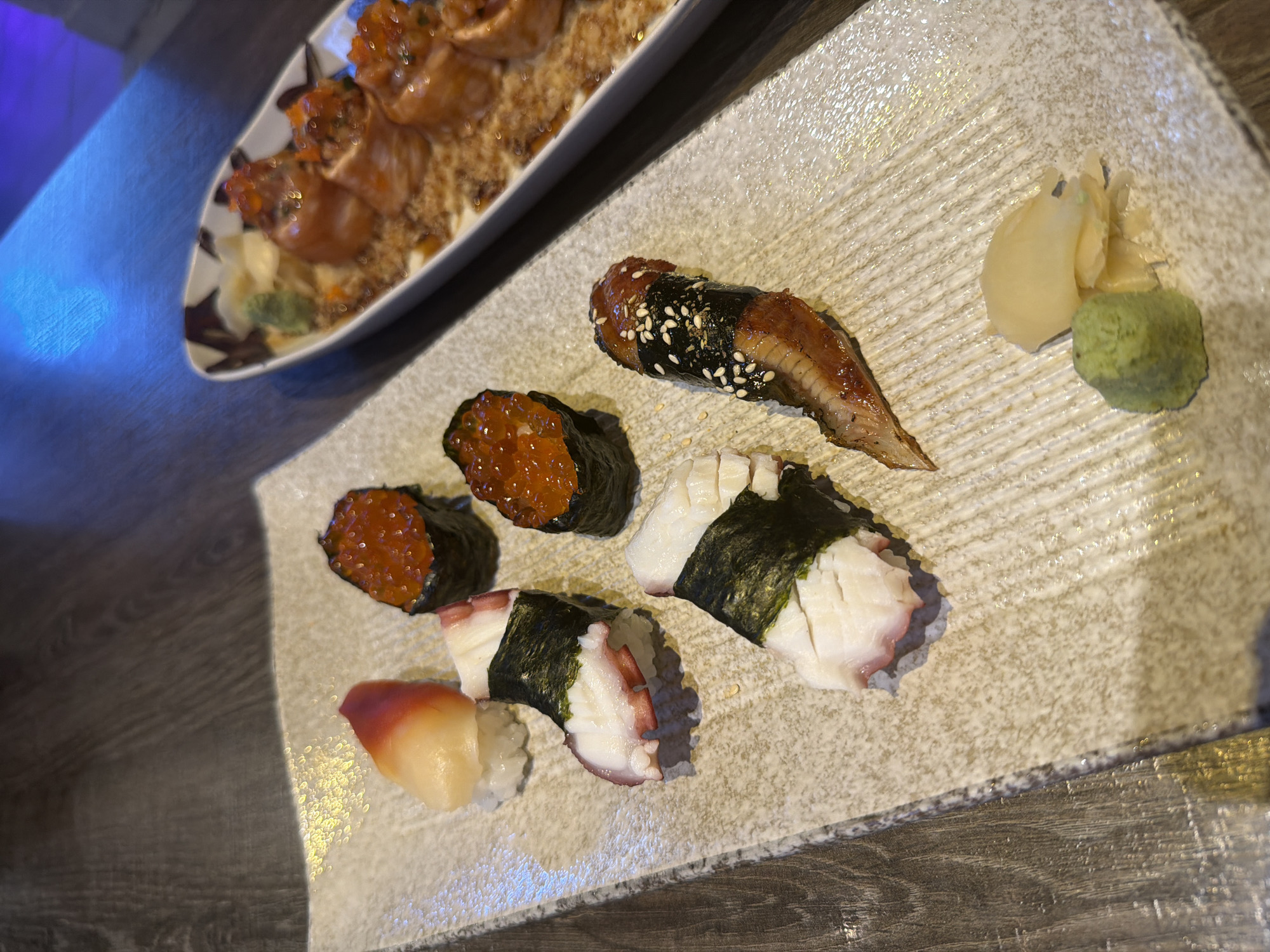The image shows a plate of sushi with several different types: ikura (salmon roe), octopus, and unagi (eel). Also visible are wasabi and pickled ginger. A second dish containing what appears to be cooked salmon with a sauce and garnish is partially visible in the upper left corner. The image appears to be taken indoors at a restaurant. It is not possible to verify the location in the city of บ้านบางรักษ์, Thailand without additional information.