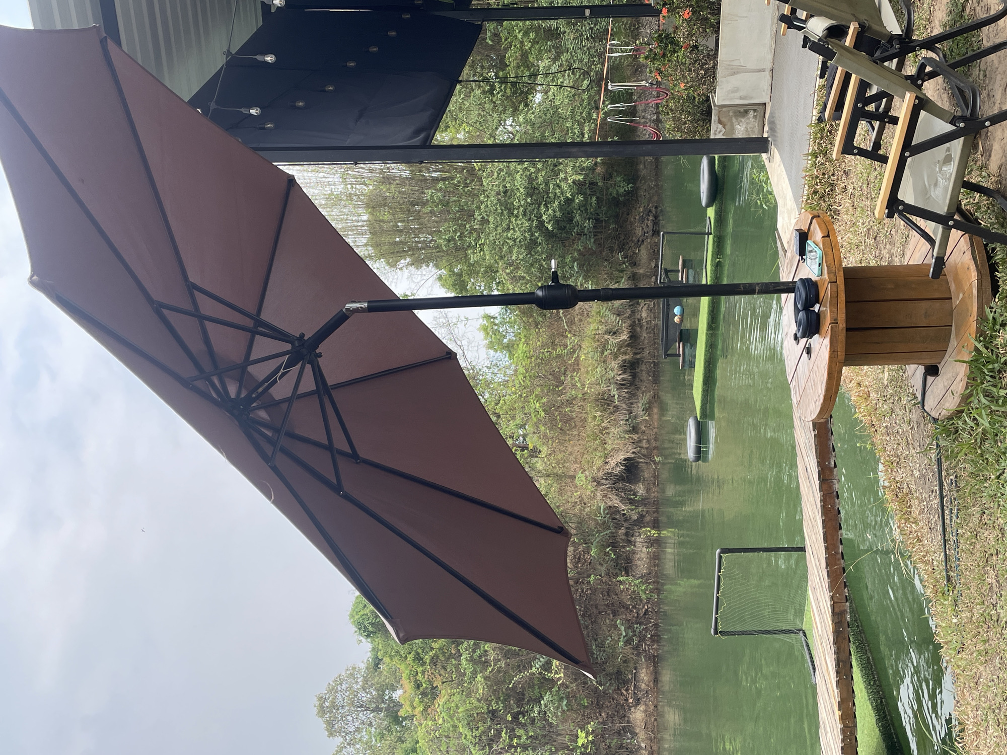 The image shows an outdoor scene at what appears to be a fishing or recreation area, potentially within Tambon Nam Phrae, Thailand. A brown umbrella is prominently featured on the left side of the image, offering shade. To the right, there is a wooden spool repurposed as a table, with items such as headphones and a phone on top. Next to the spool table, there are foldable chairs. Beyond the chairs and spool, there's a body of water with green tint, possibly a pond or lake. A wooden pathway or dock extends into the water. The landscape includes vegetation and trees in the background. Other floating objects such as tires and nets, or frame structures are in the water. The lighting suggests it's daytime, likely overcast.