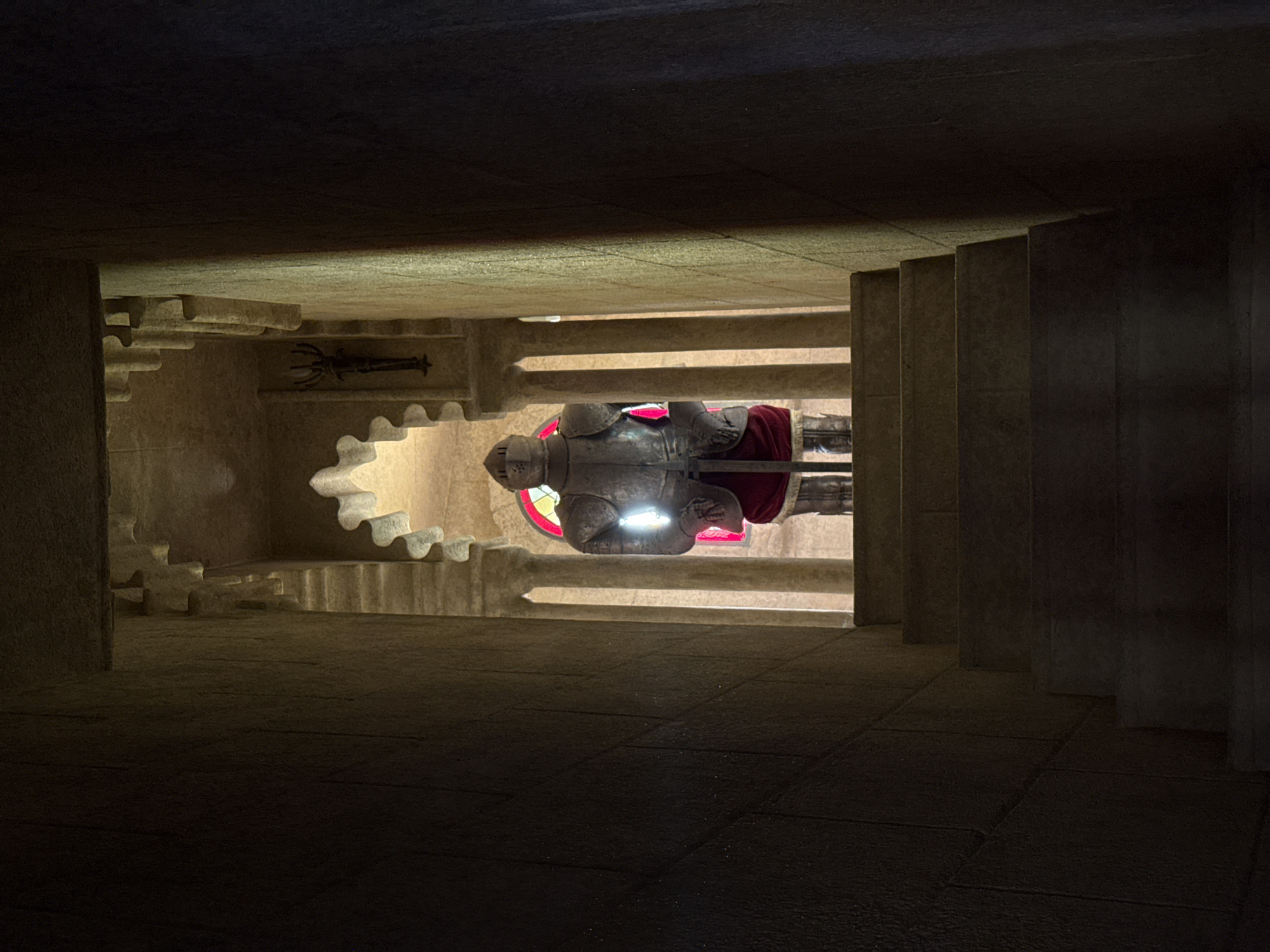 This image depicts an indoor scene, likely within a monument or historical building, given the stone architecture and the presence of a suit of armor. The setting is dimly lit, with strong shadows cast by the architecture, suggesting it is either nighttime or the interior is not well-illuminated. The primary subject is a full suit of medieval-style armor, positioned upright and seemingly on display. The armor is metallic, with visible articulation at the joints. It appears to be resting on a red cloth or cloak.
A notable feature is the reflection of the armor and its surroundings on a polished surface below, creating a mirrored image that is also visible in the frame. This reflection suggests the floor or a display base is highly reflective. In the background, behind the armor, there's a hint of a red stained-glass window, casting a colored glow. To the left of the armor, a dark, intricately shaped object, possibly a weapon or decorative artifact, is mounted on the wall. The architecture itself is characterized by stepped, carved stone elements, resembling stairs or decorative ledges. The overall mood is somber and historical, with the lighting and the subject matter evoking a sense of past grandeur or solemnity. The location context provided, Benalmádena, Spain, might suggest this is part of a local attraction or monument. No text is visible in the image.
