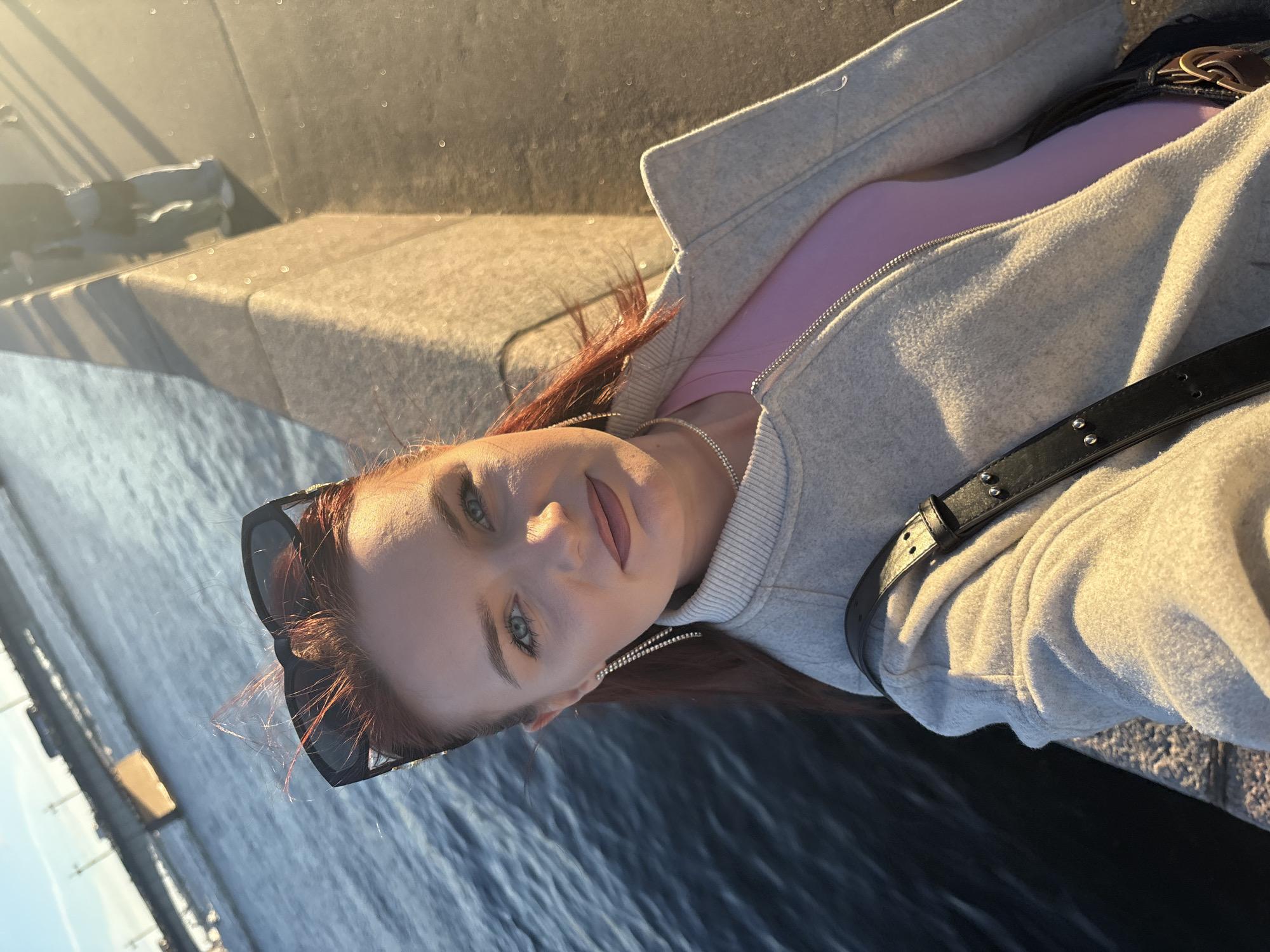 A woman with auburn hair, wearing sunglasses, a light gray jacket over a pink top, and sparkling earrings, takes a selfie outdoors. She is standing next to a stone embankment overlooking dark blue water. In the background, a bridge with cars is visible. The scene appears to be a waterfront promenade in Saint Petersburg, Russia, likely near Palace Bridge or Troitsky Bridge, given the architectural style of the bridge and the river setting. The lighting suggests it is late afternoon or early evening, with the sun casting a warm glow on the scene.