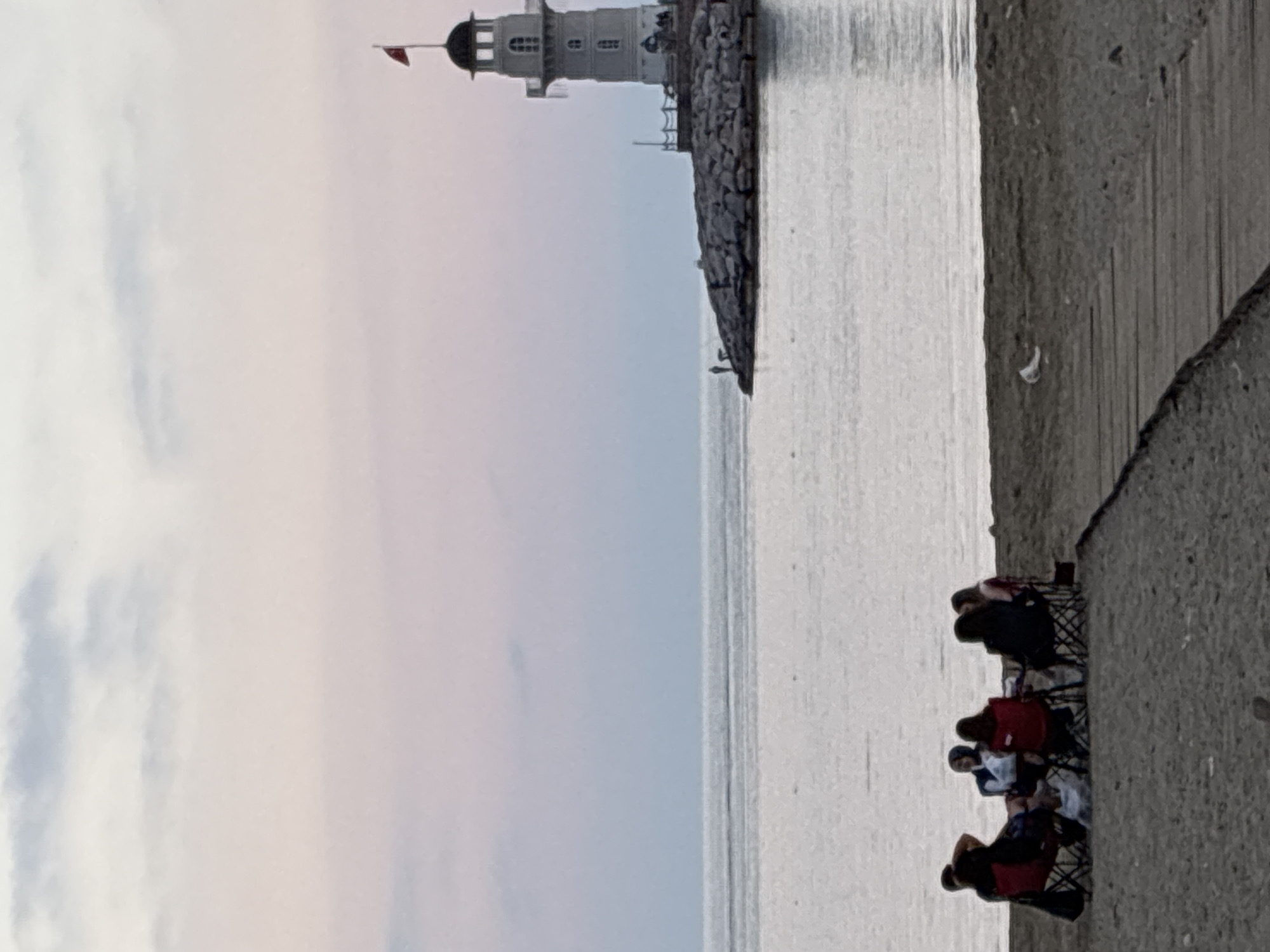 The image, oriented horizontally, presents a serene coastal scene, likely in Alanya, Turkey. On the left side, a prominent white lighthouse stands atop a rugged stone jetty or breakwater that stretches into a calm body of water. The lighthouse has a classic design with multiple windows and is crowned by a dark, rounded dome from which a small red flag flutters.
In the foreground, on the right side of the frame, a group of approximately four to five individuals are seated on portable folding chairs on a concrete or earthen bank. They appear to be relaxing, possibly enjoying the view of the sea and the lighthouse. The water surface is tranquil, reflecting the soft, diffused light from an overcast sky which occupies the upper portion of the image. The sky shows subtle cloud formations, suggesting a daytime scene without direct sunlight. The overall atmosphere conveys peace and leisure. No text is visible in the media file.