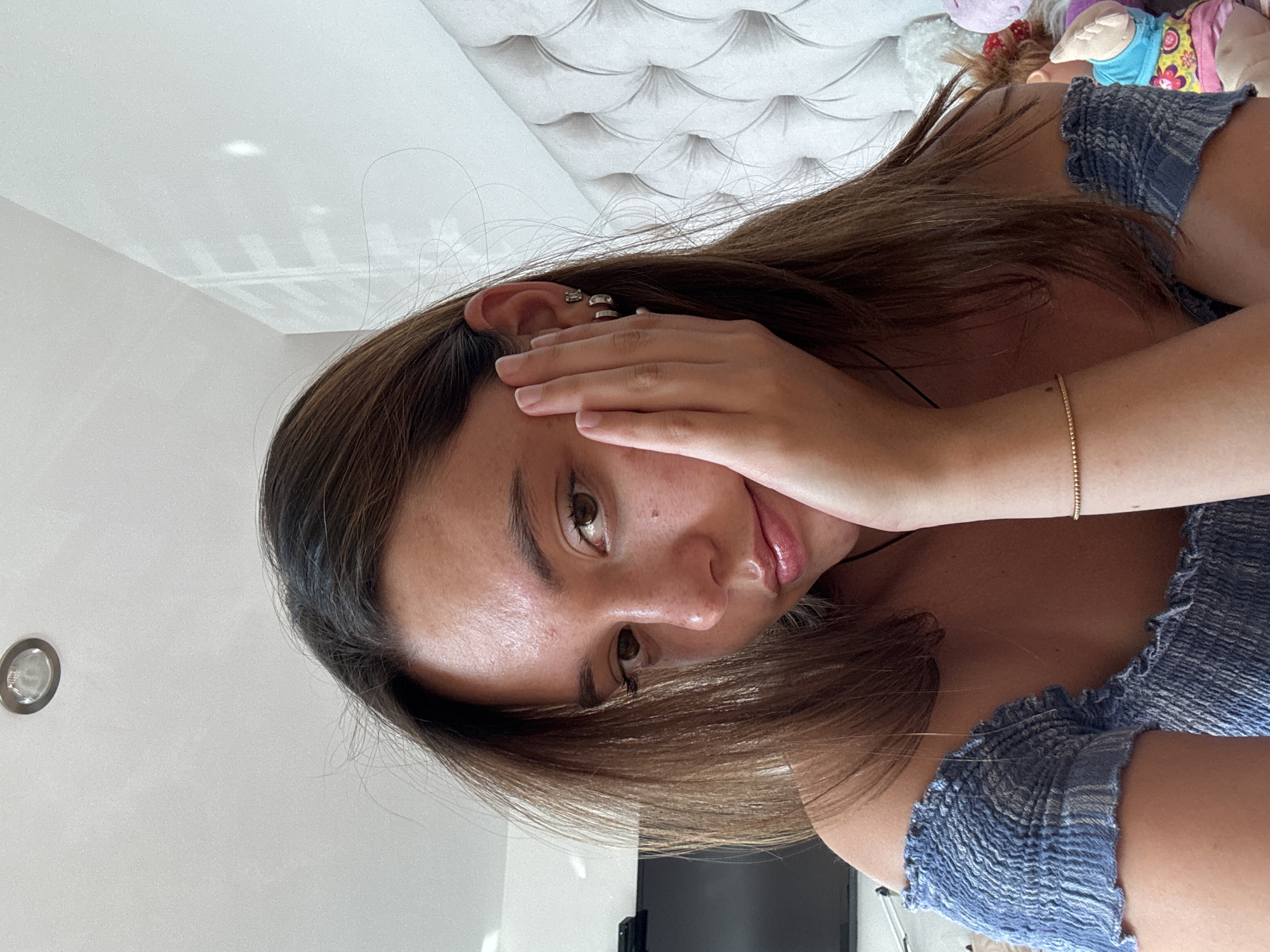 A young woman with brown hair and fair skin is seen in a close-up shot. She has her hand partially covering her face, near her forehead. Her eyes are brown, and her lips are glossy. She is wearing a blue, textured top. On her left ear, there are multiple small ear cuffs. A gold bracelet is visible on her left wrist. The background includes a white, quilted headboard and a white ceiling with a recessed light fixture. In the upper right corner, there are some blurred, colorful objects that appear to be stuffed animals or dolls. There is nothing in the image to suggest that the photo was taken in San Bernardino Tlaxcalancingo, Mexico.