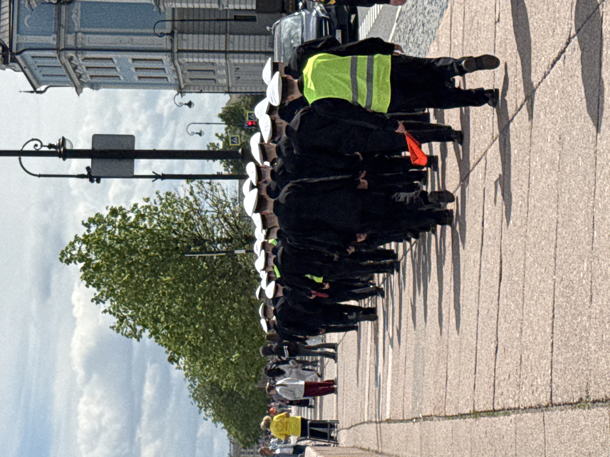 The image captures an outdoor scene on a paved urban waterfront or street, featuring a long line of uniformed individuals. The primary subjects are a procession of at least twenty people, most wearing dark, possibly navy, uniforms with distinctive white round hats, suggesting a naval or cadet formation. One person near the front of this line stands out with a bright yellow or lime green reflective safety vest over their dark uniform. Another individual in the line holds a small red flag.
The group is marching or standing in an orderly fashion from left to right across the frame, casting long shadows to their right on the light-colored pavement, indicating a sunny or partly sunny daytime. Further down the line, away from the camera, more casually dressed bystanders are visible, observing the procession.
In the background to the left, a grand, light-colored building with ornate architectural details, possibly blue or grey, lines the street. Partially visible text on the building appears to read "AIA". Above the building, a light blue sky with some soft white clouds is present. A line of lush green trees runs along the left side of the frame, separating the paved area from what might be a body of water or another part of the city. Street furniture includes black lampposts with decorative elements and a pole with a small red traffic light or signal, and an arrow sign pointing right.
The scene depicts a public event or parade in Saint Petersburg, Russia, likely during spring or summer due to the full foliage on the trees and the bright daylight. The participants appear focused, while the overall atmosphere seems to be one of quiet observation.