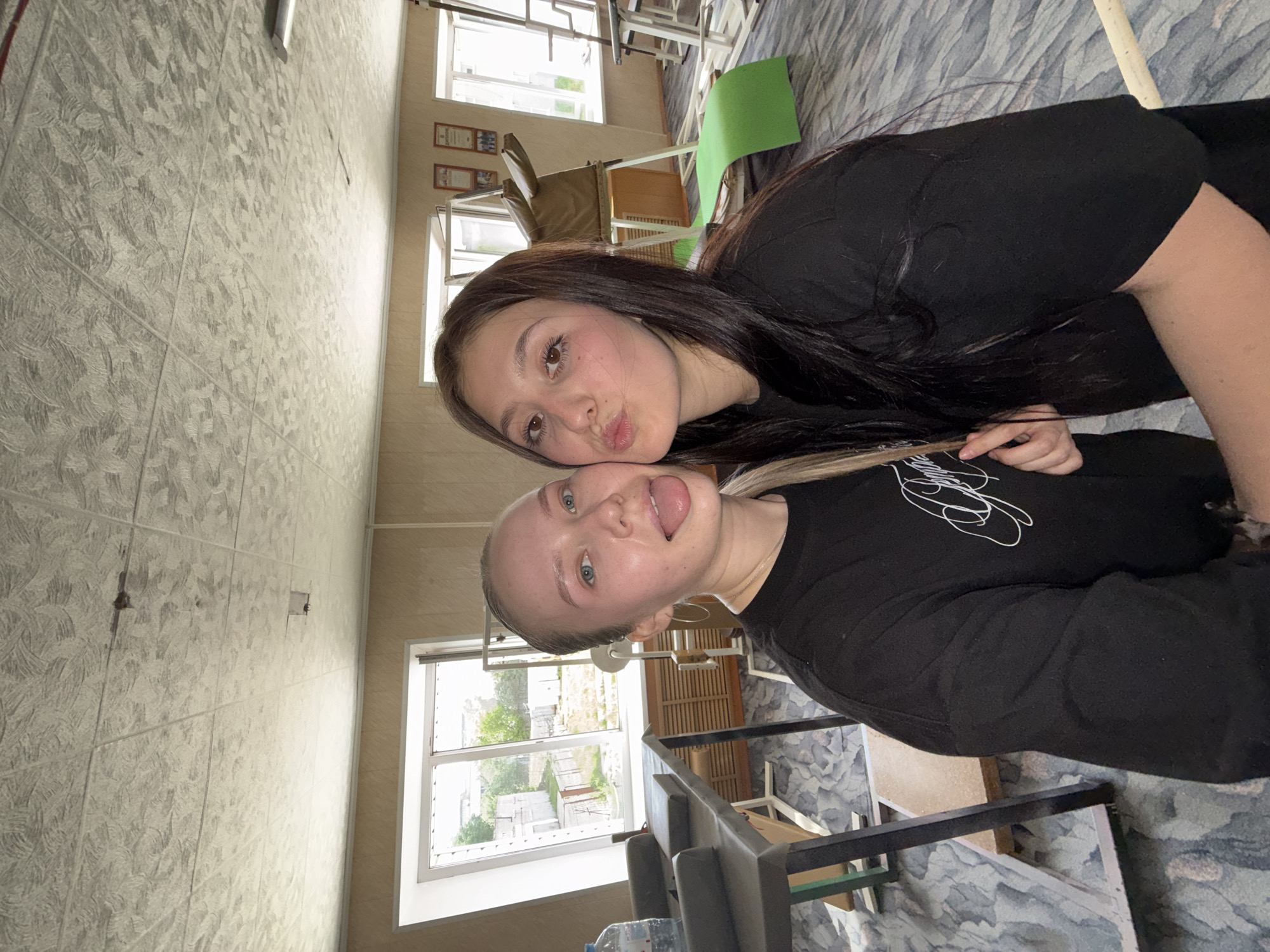 Two young women are posing closely together indoors, likely a school or public building based on the furnishings. The woman on the right has long dark hair and is making a pursed-lips face. The other woman, positioned slightly below, has blonde hair, possibly dyed, and is sticking out her tongue. Both are wearing black shirts.
The background suggests a classroom or similar setting, with desks, chairs, and windows visible. The building is located in the city of Chaikovsky, Russia, as indicated by a visible sign outside the window. The floor is covered in a patterned carpet, and the walls have tiled paneling.