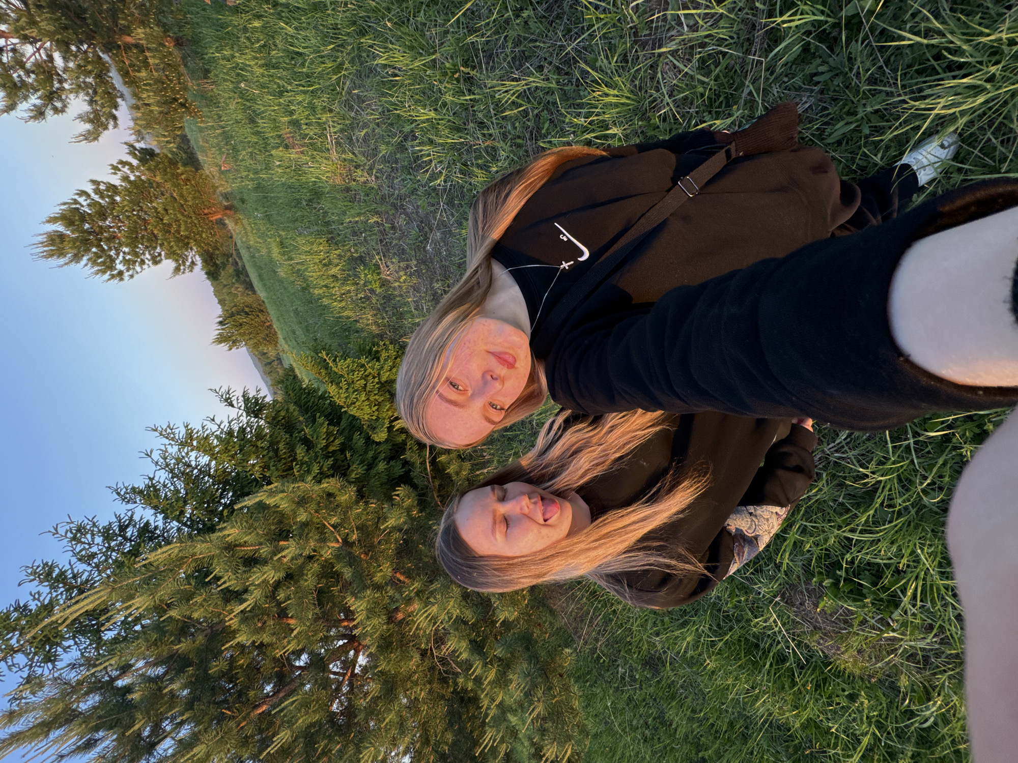 The image shows two light-skinned individuals with long blonde hair lying in grass. They are both wearing dark hoodies. The person in the foreground is sticking their tongue out. A conifer tree is visible on the left side of the frame. The photo appears to be a selfie taken outdoors during the daytime. The location is not identifiable as Zhigalki, Russia based on the visual information.