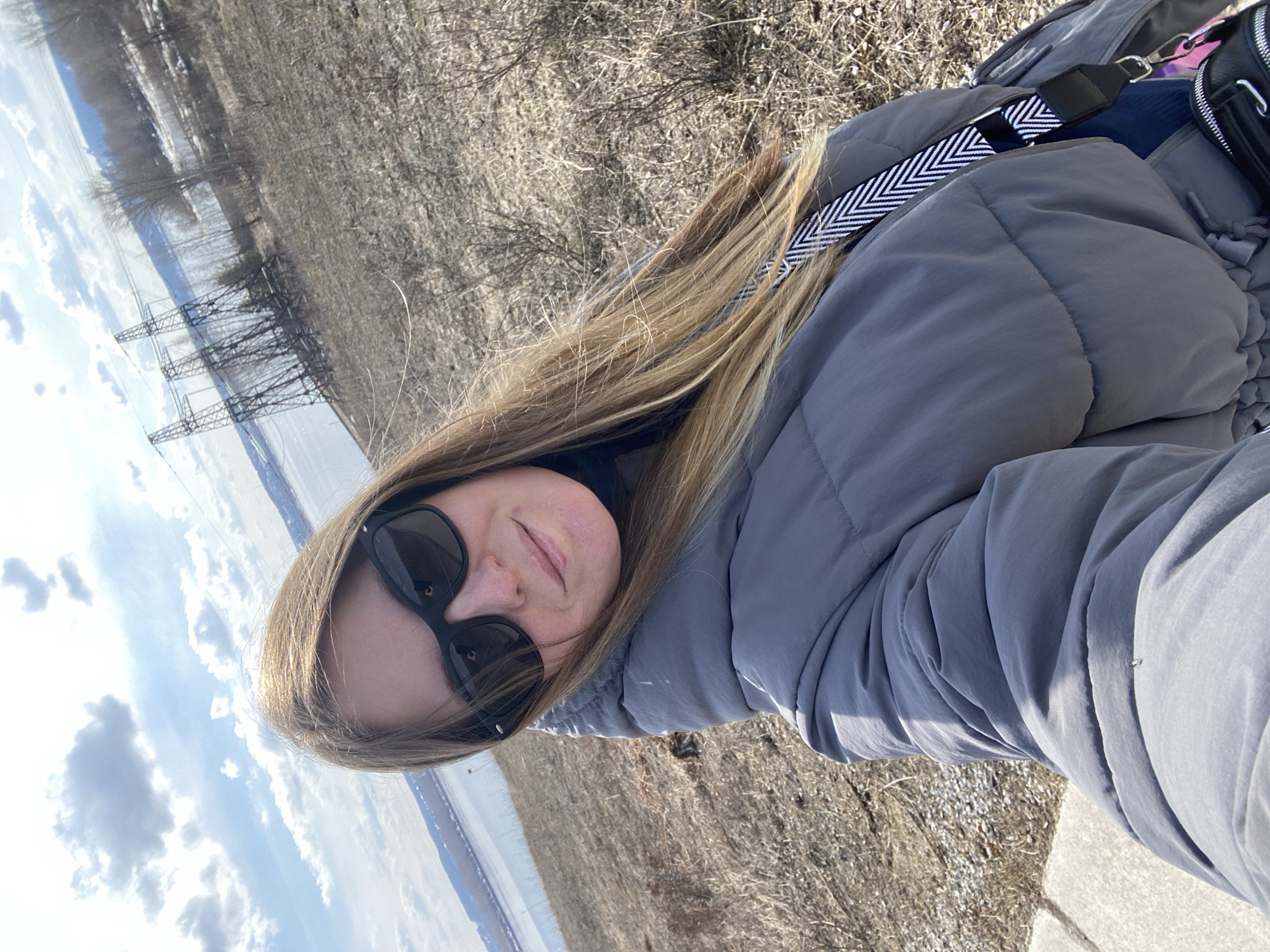 A fair-skinned person with long, blonde hair, wearing sunglasses and a grey puffer jacket, takes a selfie outdoors. The person has a small bag with a black and white strap over their shoulder. The background shows a body of water, possibly the Kama River, with a snowy bank on the opposite side. In the distance, there are power lines. The ground in the foreground is covered in dry vegetation. The location appears to be near the Kama River in the city of Chaikovsky, Russia.