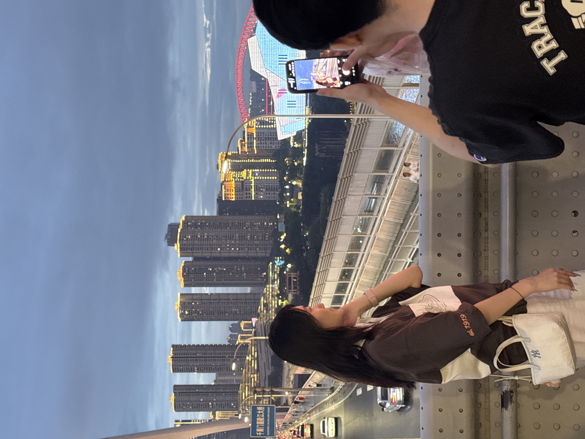 A woman with long dark hair stands on a bridge in Yuzhong District, China, looking to her left. She wears a brown and white shirt with a small white purse hanging from her left shoulder. Her right hand is on her chin. A man with short dark hair stands to her right, facing away from the camera, holding a smartphone to take a photo. The phone screen shows a picture of the woman. In the background, a city skyline with illuminated skyscrapers and a bridge with a red arch can be seen. Cars are driving on the bridge road below. The sky is cloudy and dark, indicating dusk or night.