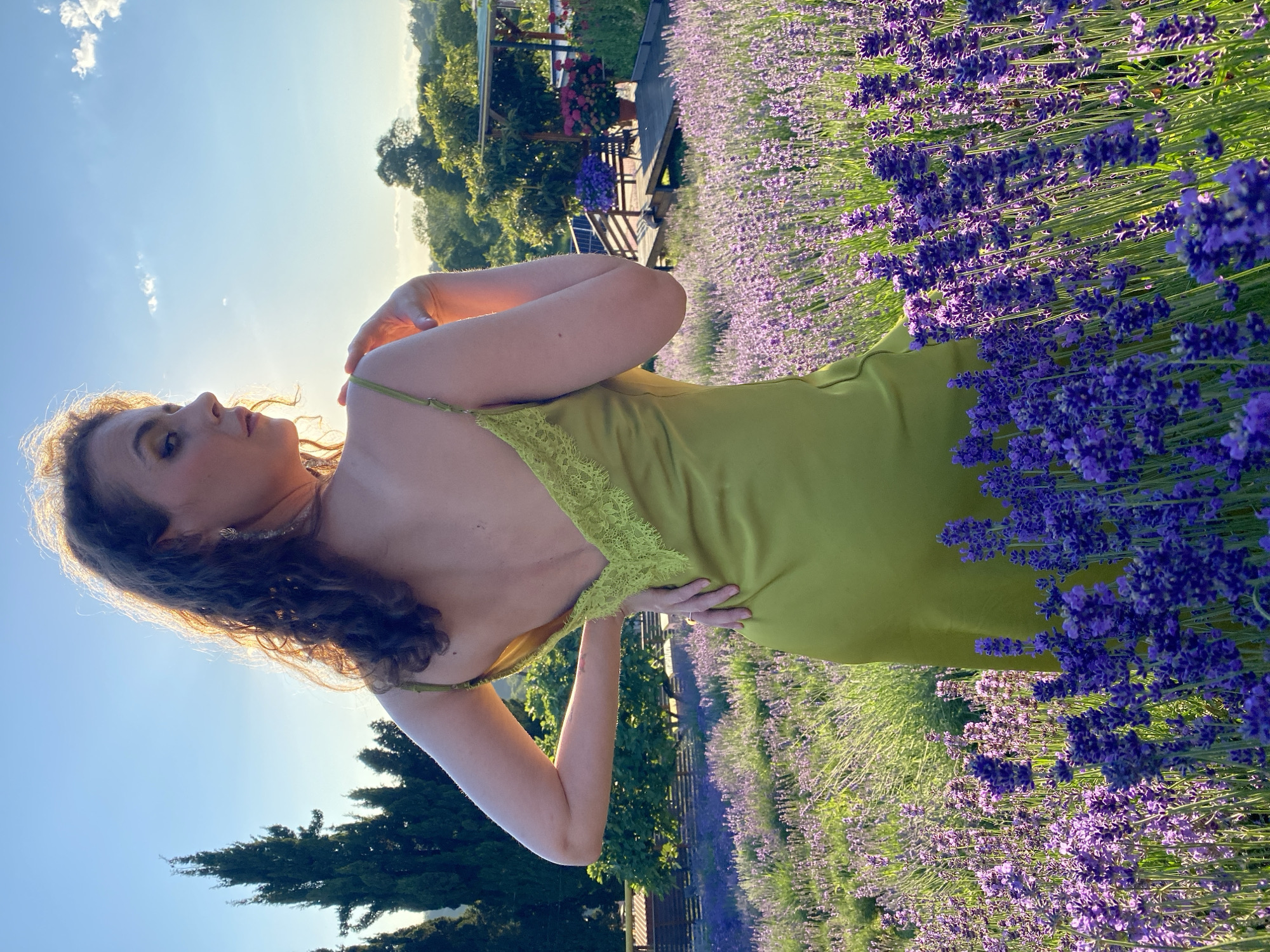 A woman with curly brown hair, wearing a lime green slip dress with lace trim, stands in a field of lavender. She is facing away from the camera, looking over her right shoulder. Her left hand is on her hip and her right hand is on her left shoulder. The sun is setting behind her, casting a warm glow. The background shows a landscape with trees and a wooden structure. This scene is located in Verkhneye Uchdere, Russia.