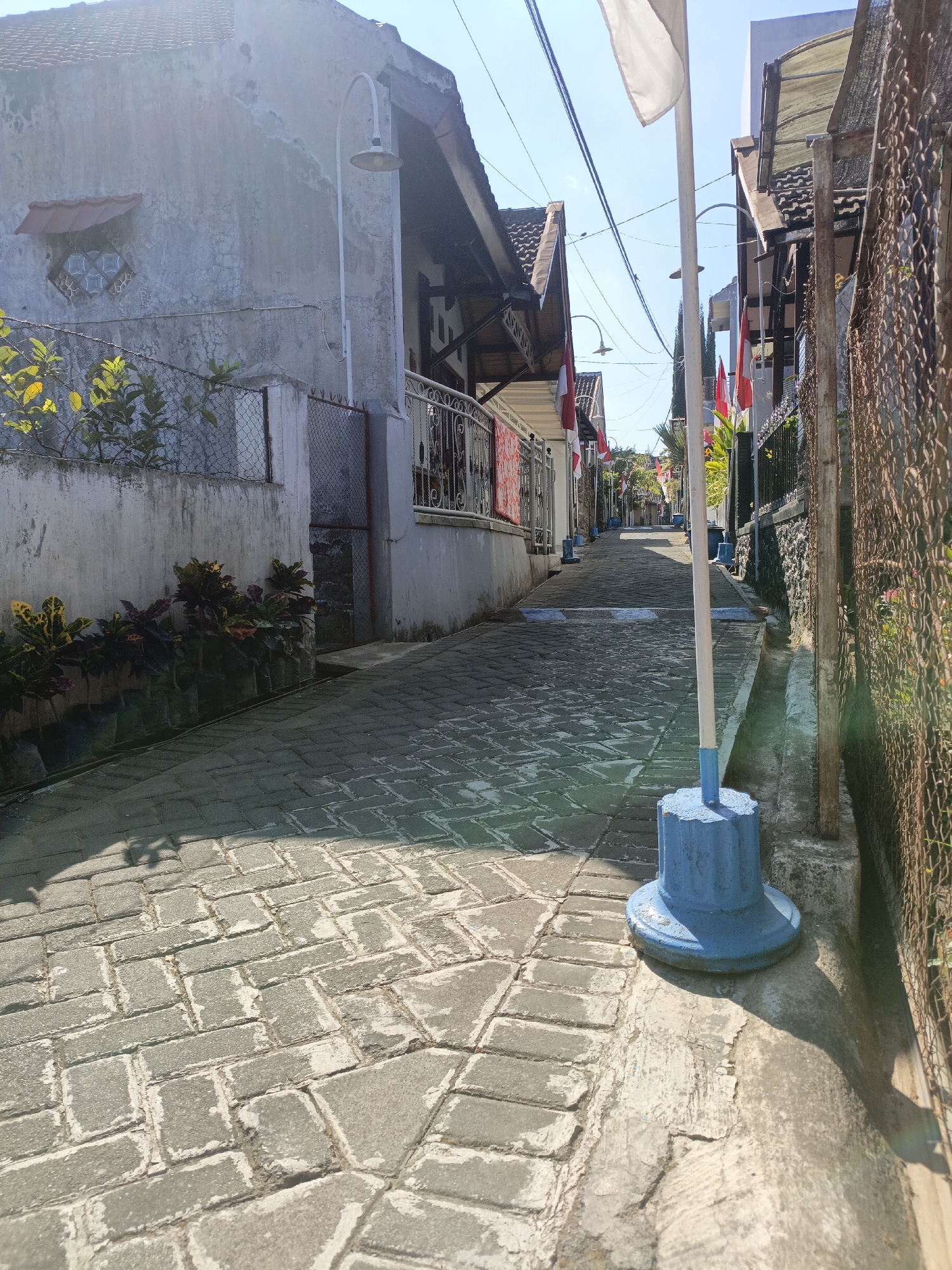 The image depicts a narrow, paved alleyway in the city of Batu, Indonesia, likely on a sunny day. The alley is flanked by residential buildings on either side. The paving stones are a grey interlocking brick pattern. Red and white flags, possibly Indonesian flags, are displayed on poles along the left side of the alley, and a white flag flies from a pole in the foreground on the right. The buildings feature tiled roofs and some have decorative metalwork. There are potted plants on the left side of the alley near the foreground. A metal fence runs along the right side of the alley. The overall impression is of a residential street that may be decorated for a national holiday or event. There are no people visible in the image.