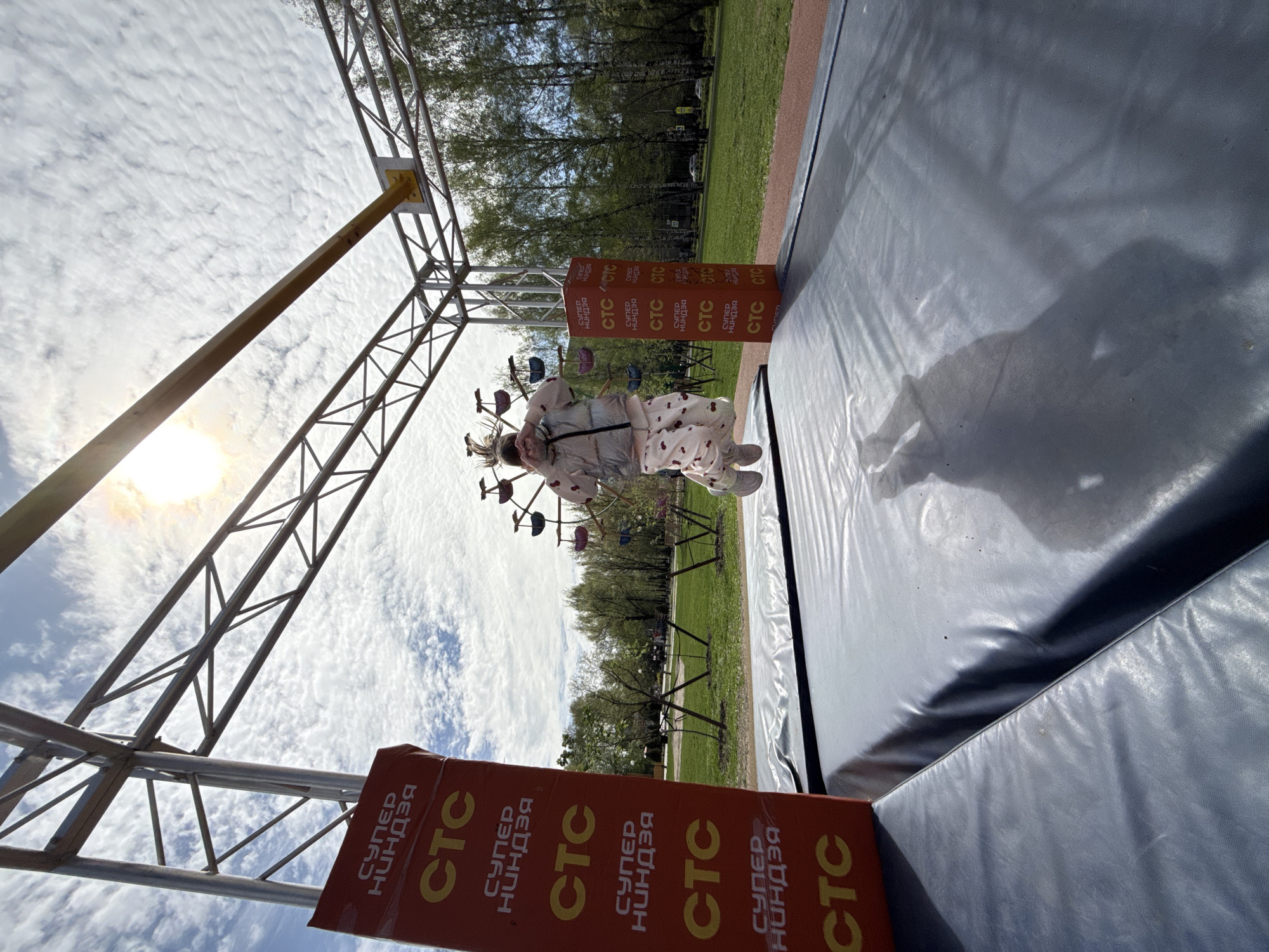 Captured at an unusual angle in what seems to be Rusanovo, Russia, the photograph features a child dressed in pajamas with cherries hanging from a monkey bar apparatus. The structure is supported by metal scaffolding with "CTC СУПЕР НИНДЗЯ" printed on orange supports. A soft, black mat rests directly under the equipment, hinting at safety precautions. The background includes green lawn, trees, and a cloudy sky with the sun shining through.