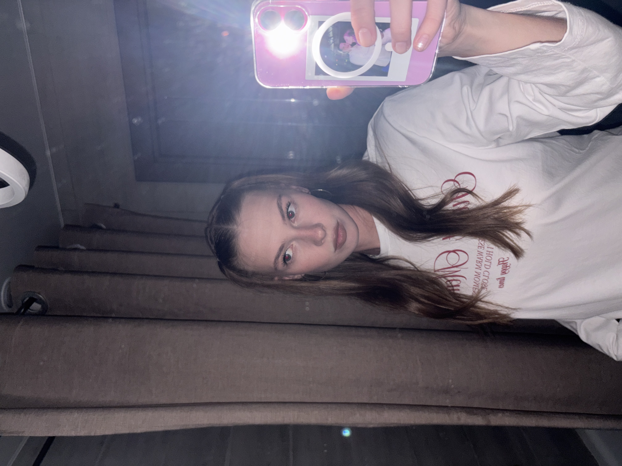 A young woman with light brown hair styled in pigtails is taking a mirror selfie in what appears to be a bathroom. She is wearing a white long-sleeved shirt with red text. She is holding a pink phone in her right hand, which displays a polaroid picture of two people. The background consists of a brown curtain and a reflective surface, likely a mirror, that shows a bright light source and what might be a door or cabinet. There are no visible elements that can definitively place this scene in the city of Perm, Russia.