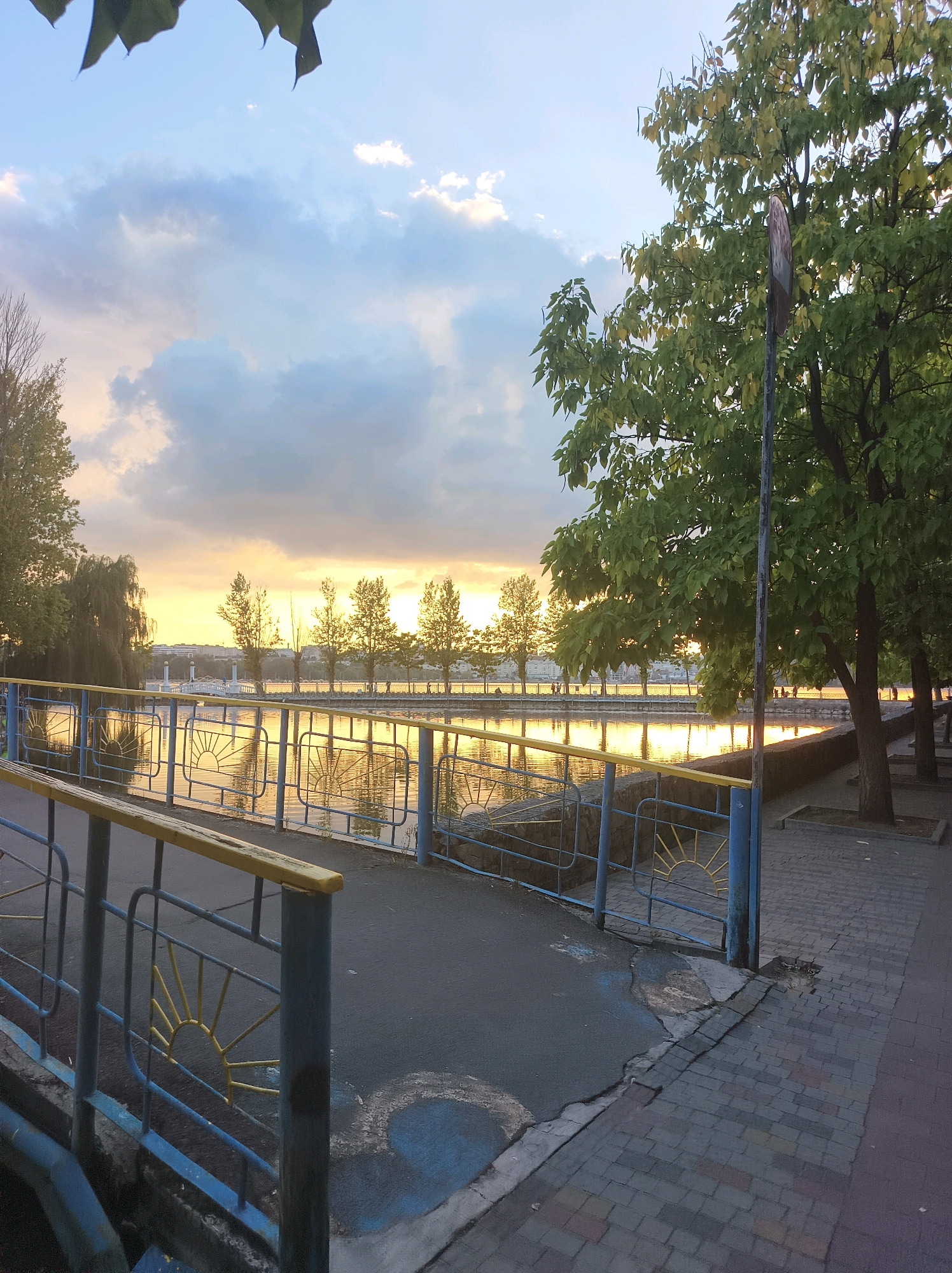 The image depicts a waterfront promenade in Ternopil, Ukraine, during either sunrise or sunset, indicated by the golden light reflecting on the calm water and the warm hues in the sky. A blue metal fence with yellow accents, featuring decorative sunburst patterns, runs along the edge of the walkway. On the right side of the frame, mature trees with lush green leaves cast shadows on the brick-paved pedestrian path. In the background, a line of trees with visible silhouettes of people walking suggests a public park or recreational area. Across the water, buildings and structures are faintly visible against the horizon. The sky is partly cloudy, with soft light filtering through. The overall scene conveys a peaceful and serene atmosphere of an urban park by the water.
