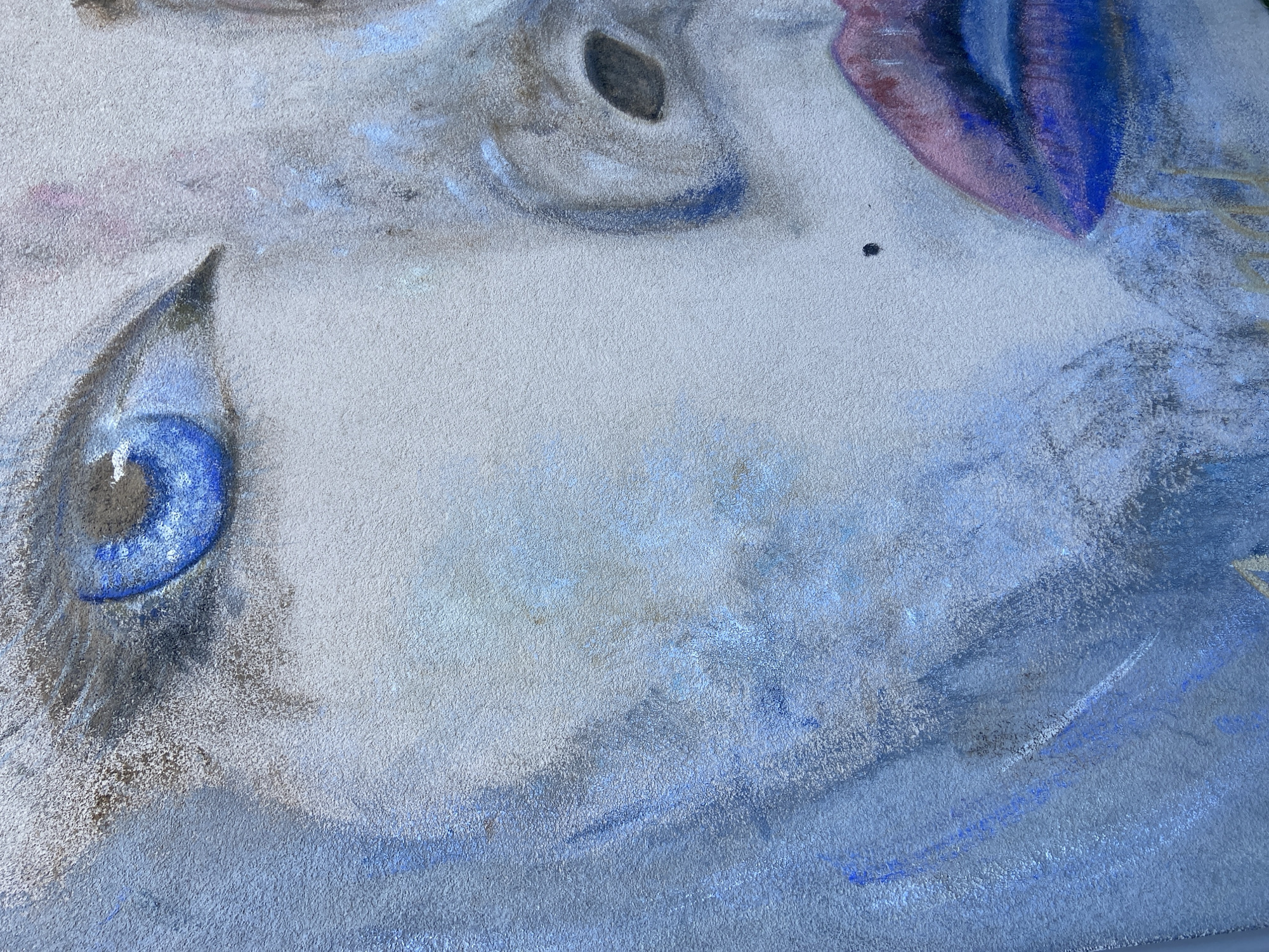 The image is a close-up of a partially completed pastel drawing, focusing on the facial features of a person. The drawing is rendered in cool tones, primarily blues, grays, purples, and whites. The eye on the left side of the image is prominent, featuring a vibrant blue iris with darker shading around it. Above this eye is an area suggestive of an eyebrow or upper eyelid, rendered in shades of gray and blue. Higher up in the image are suggestions of another eye with a dark pupil and a shape outlined in blue. To the right of the upper eye, there's a section colored with purples and blues, possibly depicting part of the face or hair. The background of the drawing is a textured, off-white surface. A small black dot is present on the upper right portion of the face, while the lower right of the frame suggests strokes of blue, giving the impression of a flowing or blurred background. The image does not feature any details related to Budva, Montenegro.