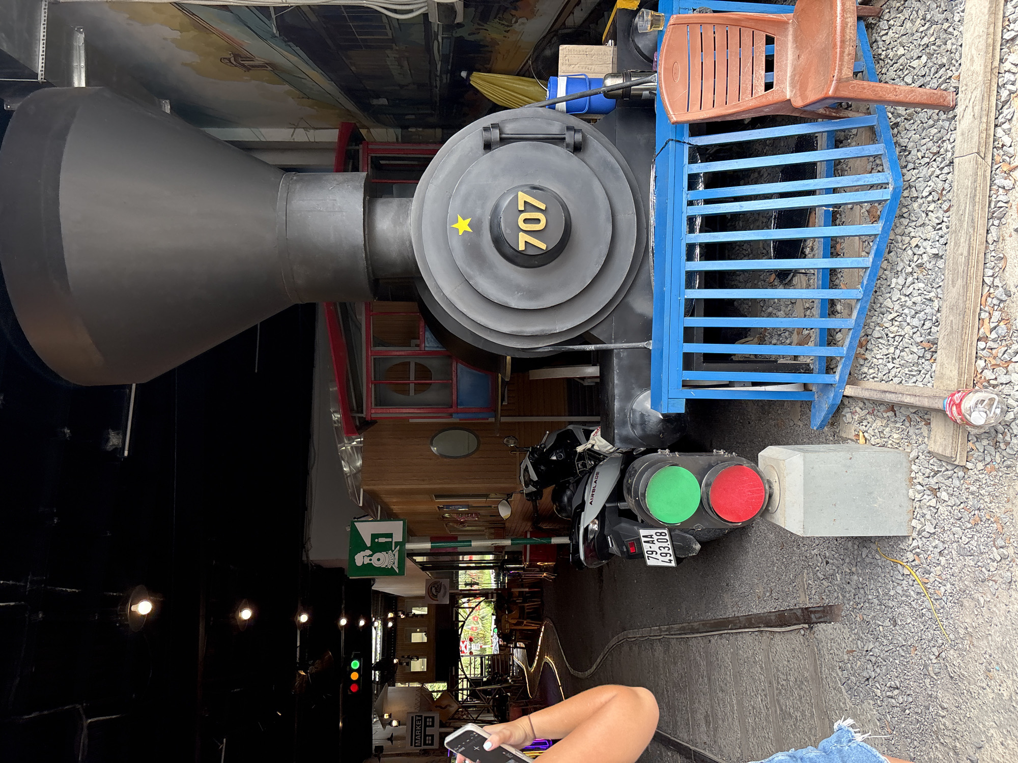 This image shows a partial view of a train engine at a location in Nha Trang Ward, Vietnam. The engine, marked with "707" and a yellow star, is black and grey with blue railings and a brown plastic chair situated on top. The engine is adjacent to a pathway that includes train tracks. A motorcycle with license plate "79-AA 493.08" is positioned in front of the engine, with a green and red traffic light-like fixture attached to its rear. To the side of the track, there's a gravel area with a grey concrete block. In the background, there is a partially visible building and a market sign. A person is holding a smartphone in the foreground. The photo is taken indoors and has been rotated counterclockwise.