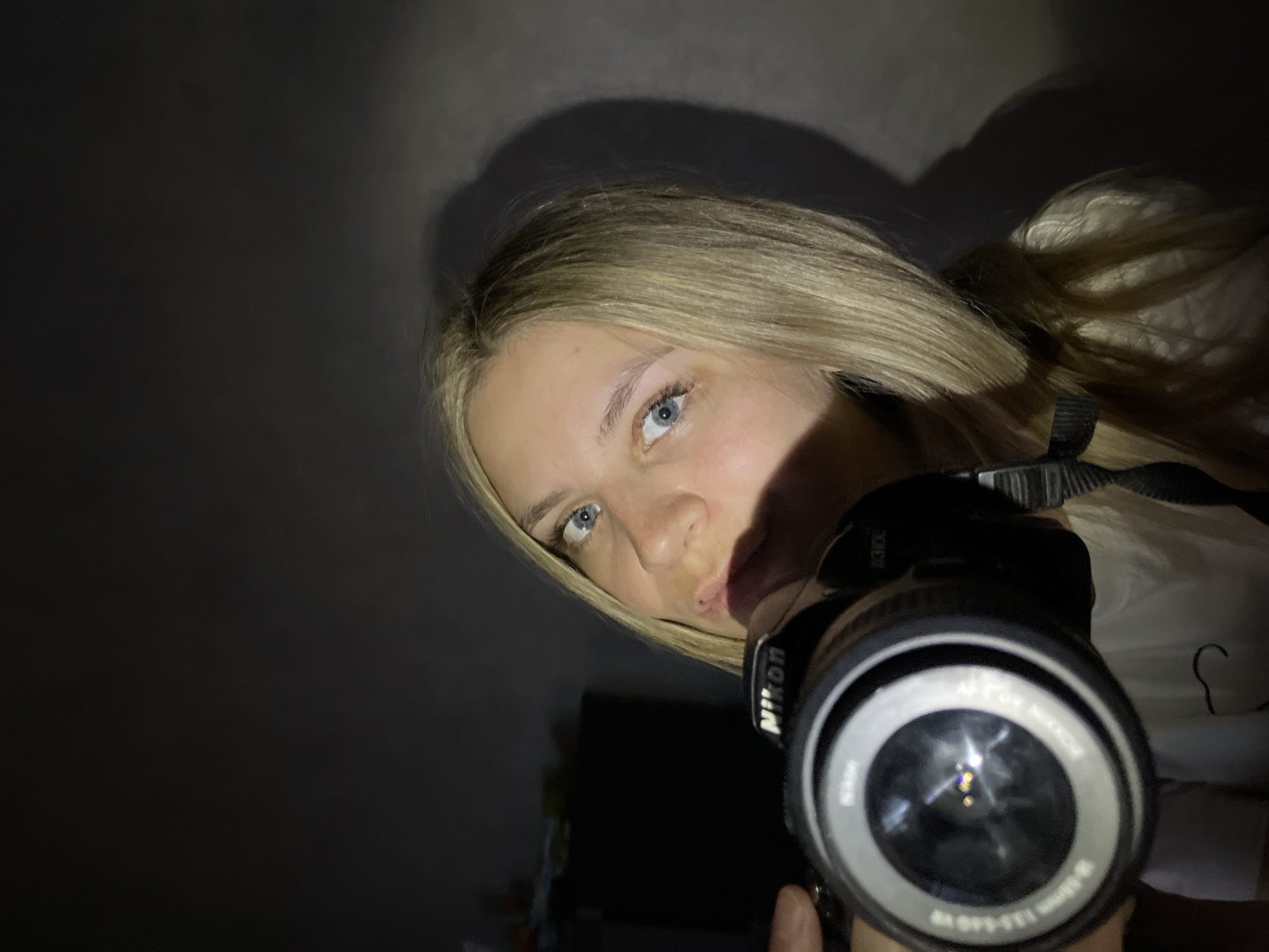 A person with light blonde hair and blue eyes is holding a Nikon camera and taking a selfie. The environment is dimly lit, with a dark background suggesting an indoor location. A shadow is cast on the wall behind the person. The camera lens is prominently featured in the foreground. The city of Chaikovsky, Russia, is not visibly apparent in this image.
