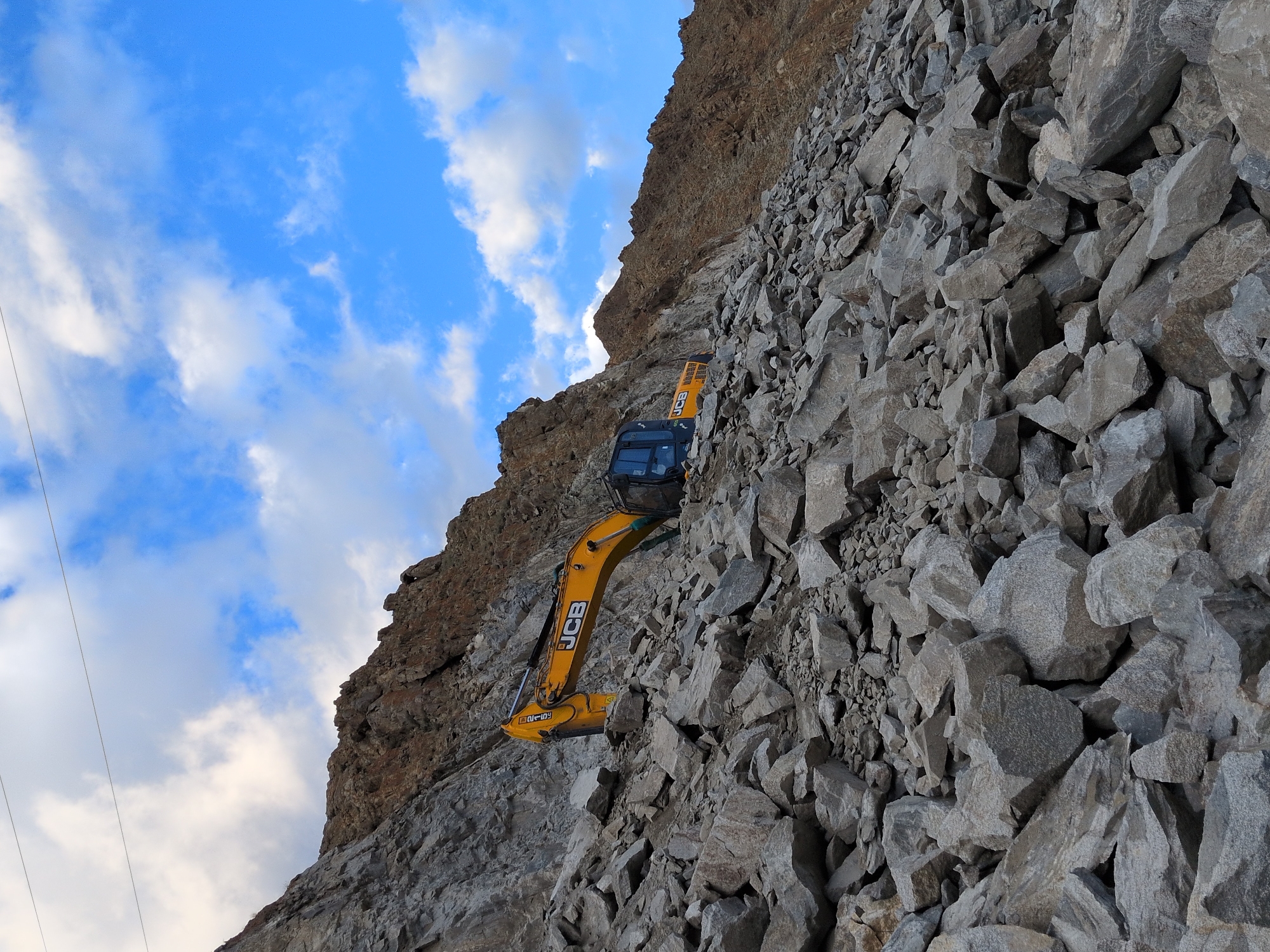 A yellow JCB excavator is positioned on a steep, rocky slope in Leh, India, amidst a large pile of grey, angular stones. The excavator's arm is extended, and its bucket is not visible. The machine has the "JCB" logo clearly displayed on its boom. A portion of the excavator's black cabin is visible. The background consists of a rugged, brown mountain face and a bright blue sky with scattered white clouds. Power lines are visible in the upper left quadrant of the image. The scene appears to depict a construction or quarrying operation in a mountainous, arid environment.
