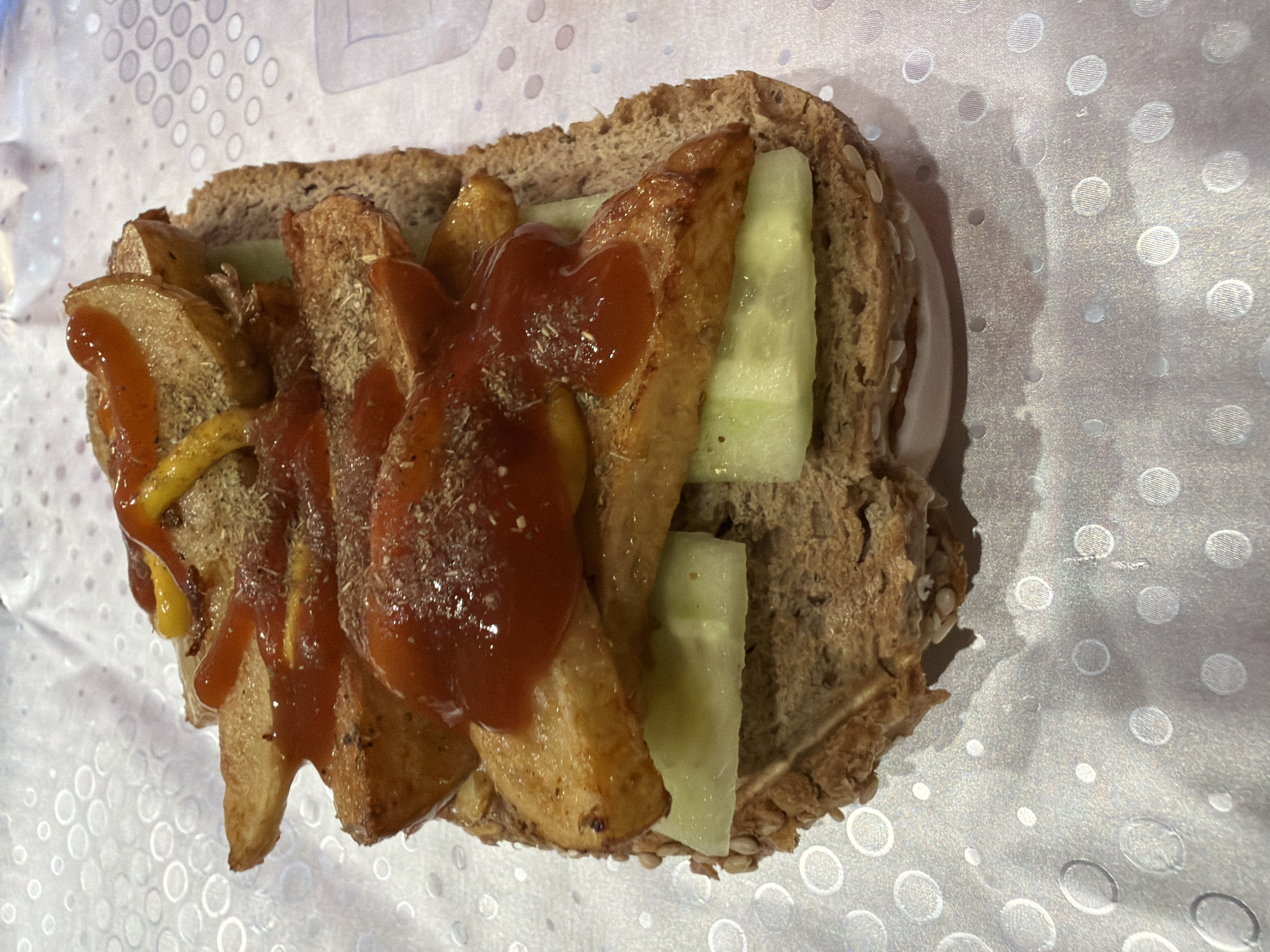 A sandwich is shown on what appears to be a patterned paper. The sandwich is made with whole wheat bread, and is filled with french fries, cucumber slices, ketchup, and mustard. It's not possible to tell if this is in Varna, Bulgaria.