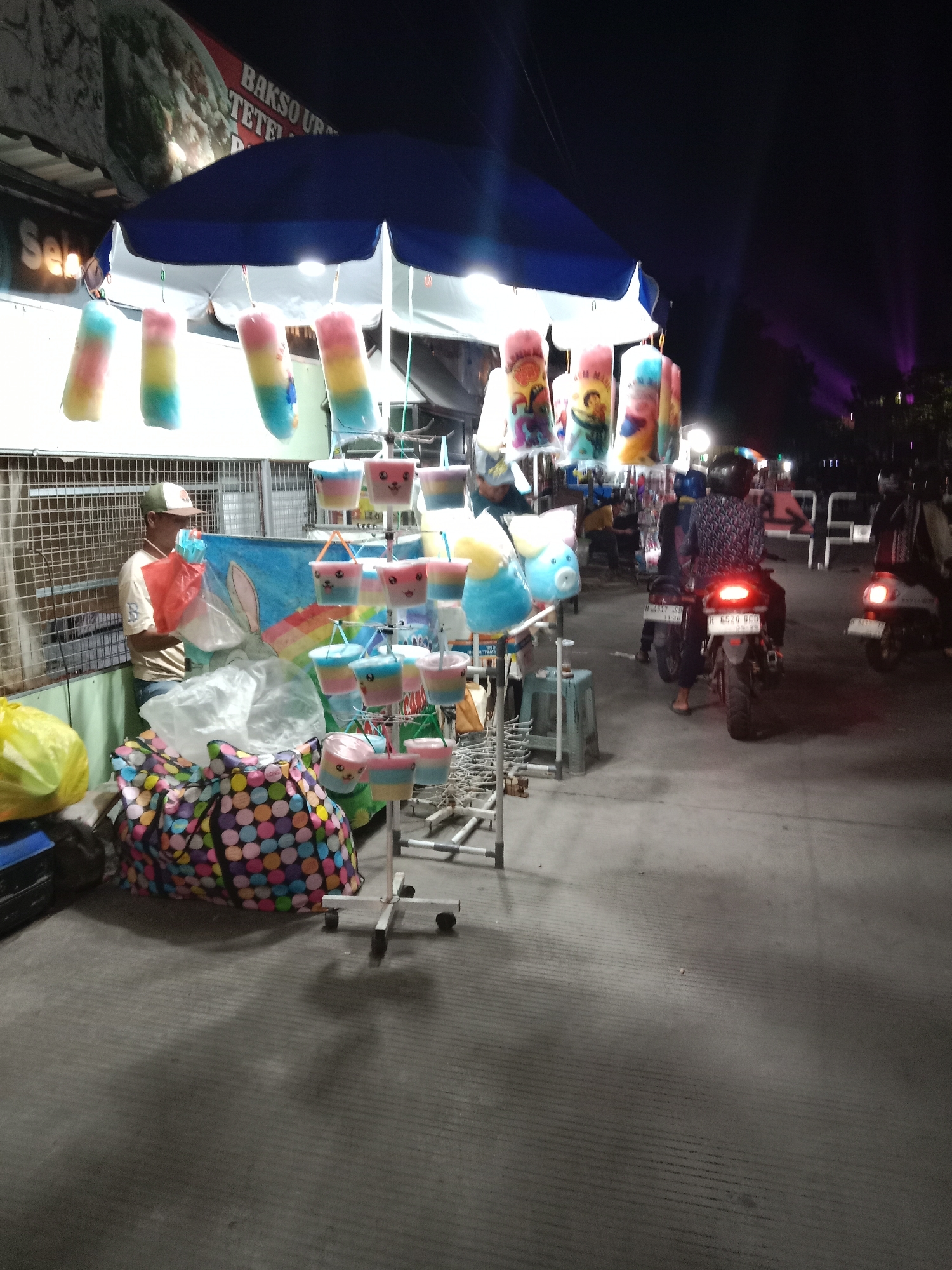 The image captures a nighttime scene on a street in Kebondalem, Indonesia, likely a market or roadside vendor area. The foreground features a stall with a blue and white striped canopy, displaying numerous colorful cotton candy treats in plastic packaging hanging from racks. These treats are predominantly pink, yellow, and blue. To the left, a person wearing a light-colored t-shirt and a baseball cap stands behind a counter, holding a red plastic bag and looking at a large, colorful polka-dotted bag on the ground. A yellow plastic bag is also visible to the left. In the background, several motorcycles are parked or being ridden, with their taillights illuminated. One motorcycle in the center has a rider wearing a patterned shirt, and another to the right has a rider wearing a dark jacket. The street surface appears to be paved. The background shows dimly lit buildings and trees, with faint purple lights emanating from further down the street, suggesting a festival or event. A sign with Indonesian text, including "BAKSO UR" and "TETEL," is visible above the canopy.
