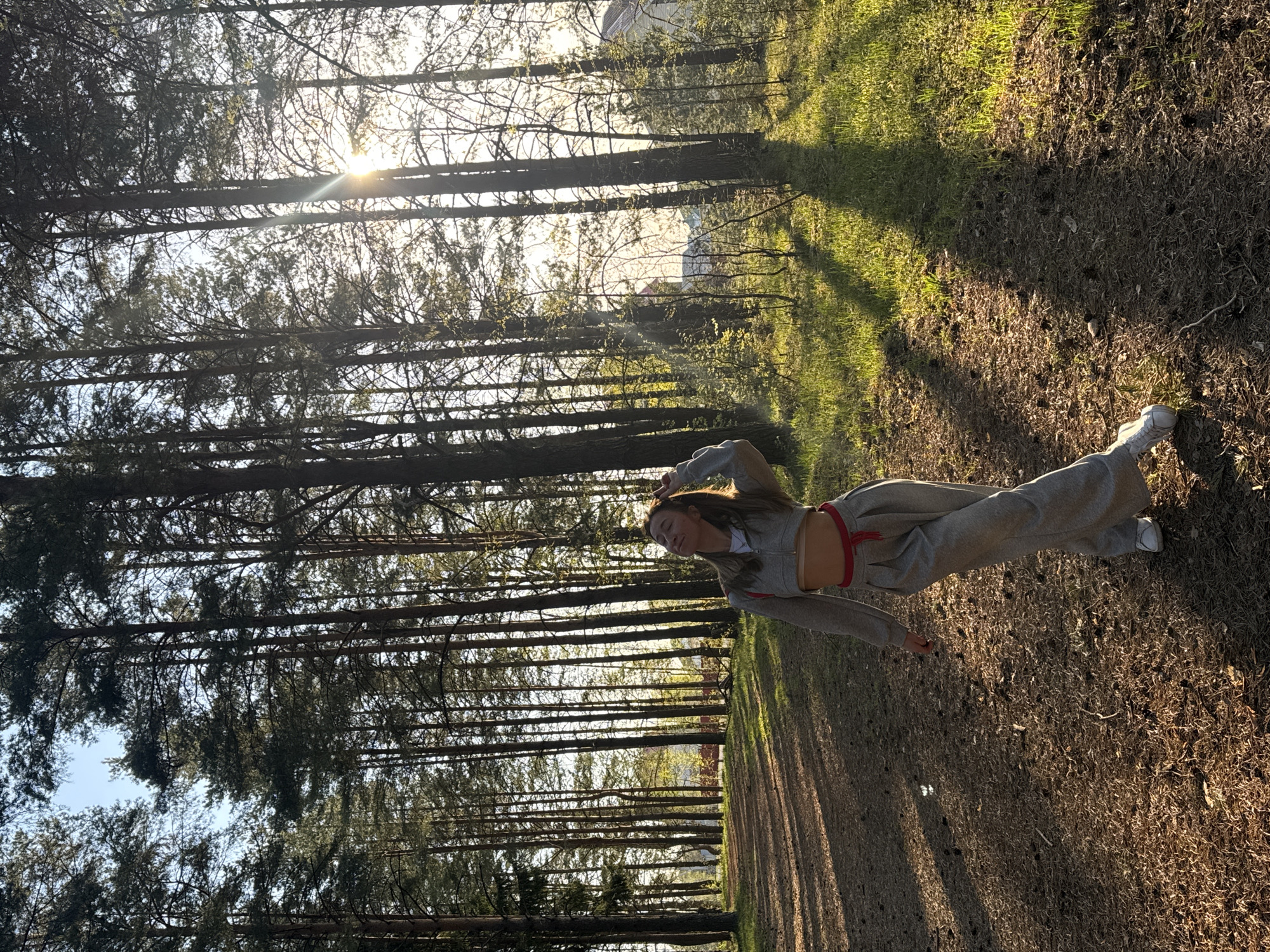 A woman stands in a forest in Прикамский, Russia, facing the viewer. She is wearing a gray sweatsuit with a red belt and white sneakers. The sun is shining through the trees. The ground is covered in dry leaves and grass. There is a building visible in the background.