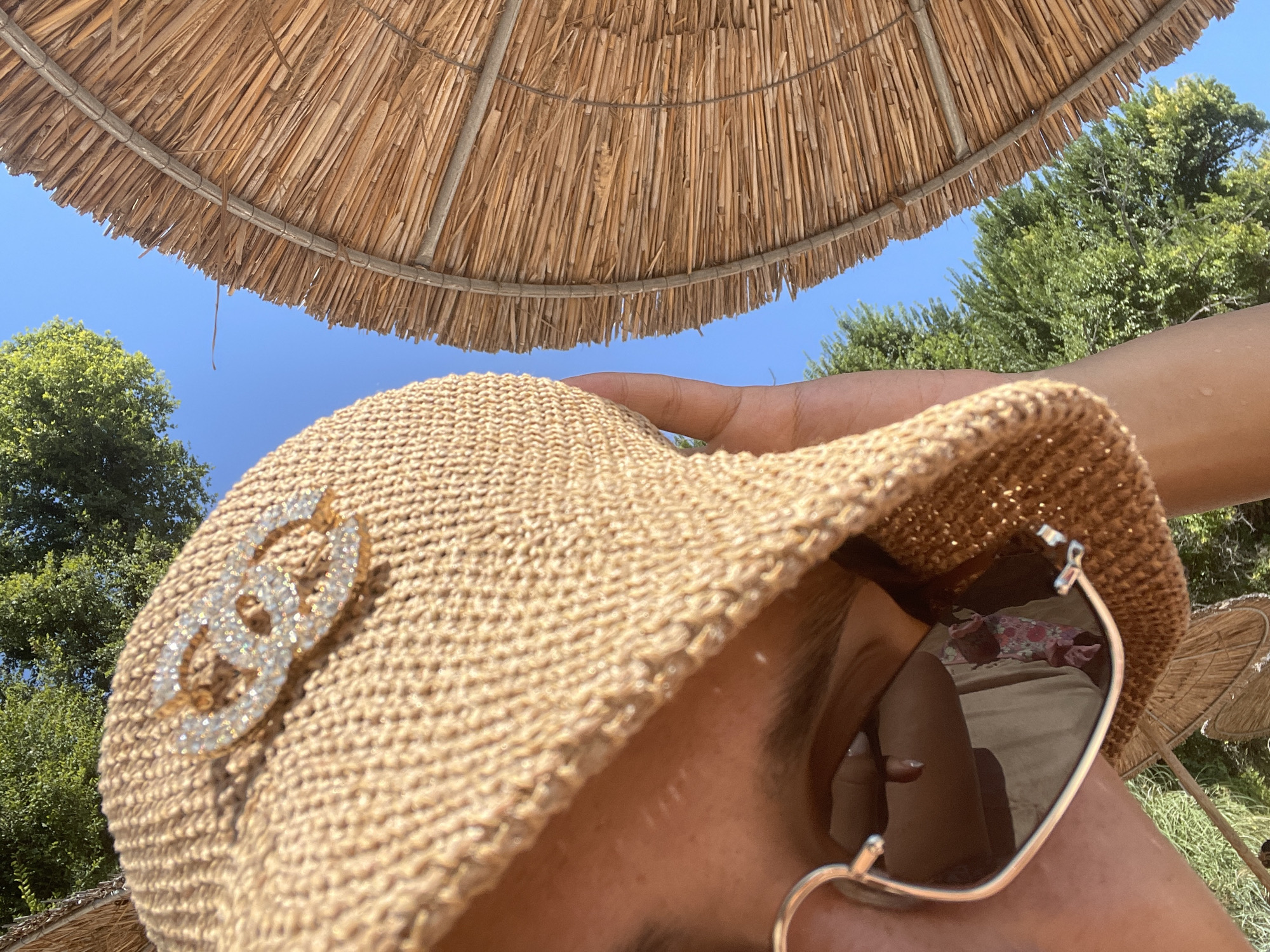 In Chernomorets, Bulgaria, a person wearing a straw hat with a diamond-encrusted logo and heart-shaped sunglasses is looking downwards. The person is partially obscured by the hat and sunglasses, but their eye and forehead are visible. A straw umbrella is visible on the right side of the image, and green trees and a blue sky are in the background. The image appears to be taken on a sunny day.