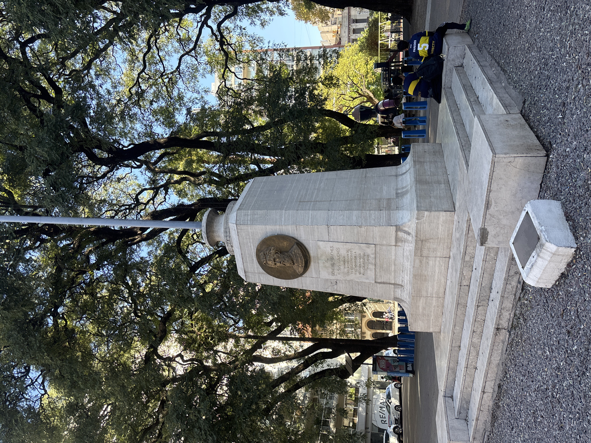 An outdoor scene captures a monument and its surroundings in Buenos Aires, Argentina, under bright daylight.
The central subject is a tall, light-colored stone monument, likely made of marble or granite, with a stepped base. A flagpole extends upwards from its top, though the flag itself is not visible. Embedded on one side of the monument is a bronze medallion featuring the bust of a man, accompanied by a rectangular plaque. The plaque's inscription reads: "AL GENERAL GUEMES Y SUS GAUCHOS," followed by "HOMENAJE DE LA COMISION DE FESTEJOS PATRIOS Y VECINDARIO DE LA SECCION 21 DE LA POLICIA FEDERAL," and dated "17-VI-1966."
Behind the monument, mature trees with dense, green foliage fill the upper and left portions of the frame, their branches forming intricate patterns against a clear blue sky. Their shadows suggest a sunny day, possibly morning or early afternoon. Below the trees, several individuals are seated on blue plastic stools or benches. One person is prominently wearing a blue and yellow soccer jersey with the name "PAREDES" and the number "5," along with the sponsor "DIRECTV." Another person is partially visible near them.
The ground around the monument and benches is covered in small grey gravel. In the background, beyond the trees and people, several multi-story buildings characteristic of an urban environment are visible, along with parked cars, including a white van with "RE/MAX" branding. A small yellow sign with "ZERZ" is also partially visible. The scene depicts a common public space, likely a park or plaza, with people casually resting or gathering near the historic monument.