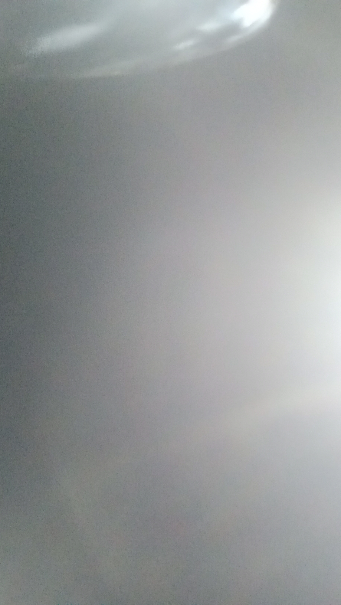 The image displays a close-up, out-of-focus view of what appears to be a gray, slightly textured surface, possibly a wall or ceiling. There are areas of bright light on the upper portion of the image, suggesting a light source above and to the right. The lighting creates a gradient, with the left side of the image being darker and the right side gradually becoming brighter and washed out by the light. There are subtle variations in tone and texture, but no distinct objects or figures are discernible. The overall impression is one of a plain, illuminated surface. The image provides no specific details that can be used to locate it in Kuta, Indonesia.