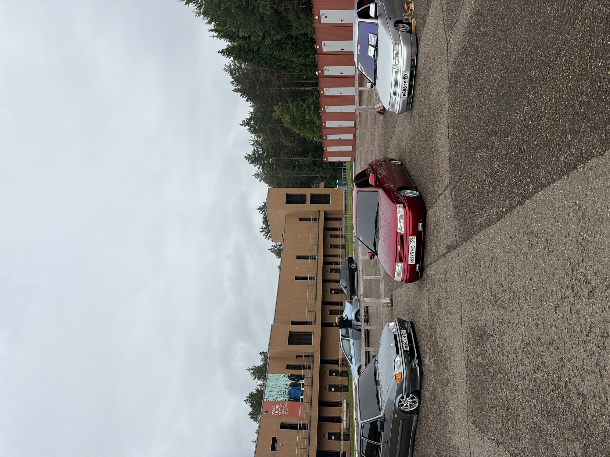 The image depicts an outdoor scene in Прикамский, Perm Krai, Russia, under an overcast sky. The foreground is a paved asphalt surface with visible cracks and wear. Three heavily modified Lada sedans are prominently parked in a triangular formation. The grey Lada Samara 2 (VAZ-2115) on the left features a low stance, aftermarket multi-spoke wheels, and tinted windows, displaying license plate "M680KM 159". A red Lada 110-series sedan, also with a low stance and tinted windows, is positioned in the middle, showing license plate "M879MO 159". On the right, a silver Lada 110-series sedan exhibits similar modifications, including a low stance and tinted windows, with a white sign visible on its windshield and license plate "P686TE 159".
In the middle ground, a male individual in a dark jacket stands beside a light blue Lada 110-series sedan, interacting with its rear door or trunk area. A dark Lada sedan is partially visible parked further behind the red structures. Behind these vehicles, a modern, multi-story building with a brown/wood-paneled facade and dark-framed windows spans the background. A large red banner on the building reads "НОВЫЙ ВЗГЛЯД НА СПОРТ / ОДЕЖДА • ОБУВЬ • СПОРТПИТ • ПАЛКИ" (NEW LOOK AT SPORT / CLOTHING • FOOTWEAR • SPORTS NUTRITION • SKI POLES) and displays an image of four people. To the right of the building, a row of approximately nine uniform, red modular structures, possibly storage units or changing rooms, each with a white door and vertical white stripe, are visible. A dense coniferous forest line forms the horizon behind the building and structures, indicating a natural, tree-lined environment surrounding the developed area. Wooden fence posts are also visible in the middle ground, separating the paved area from the red structures.