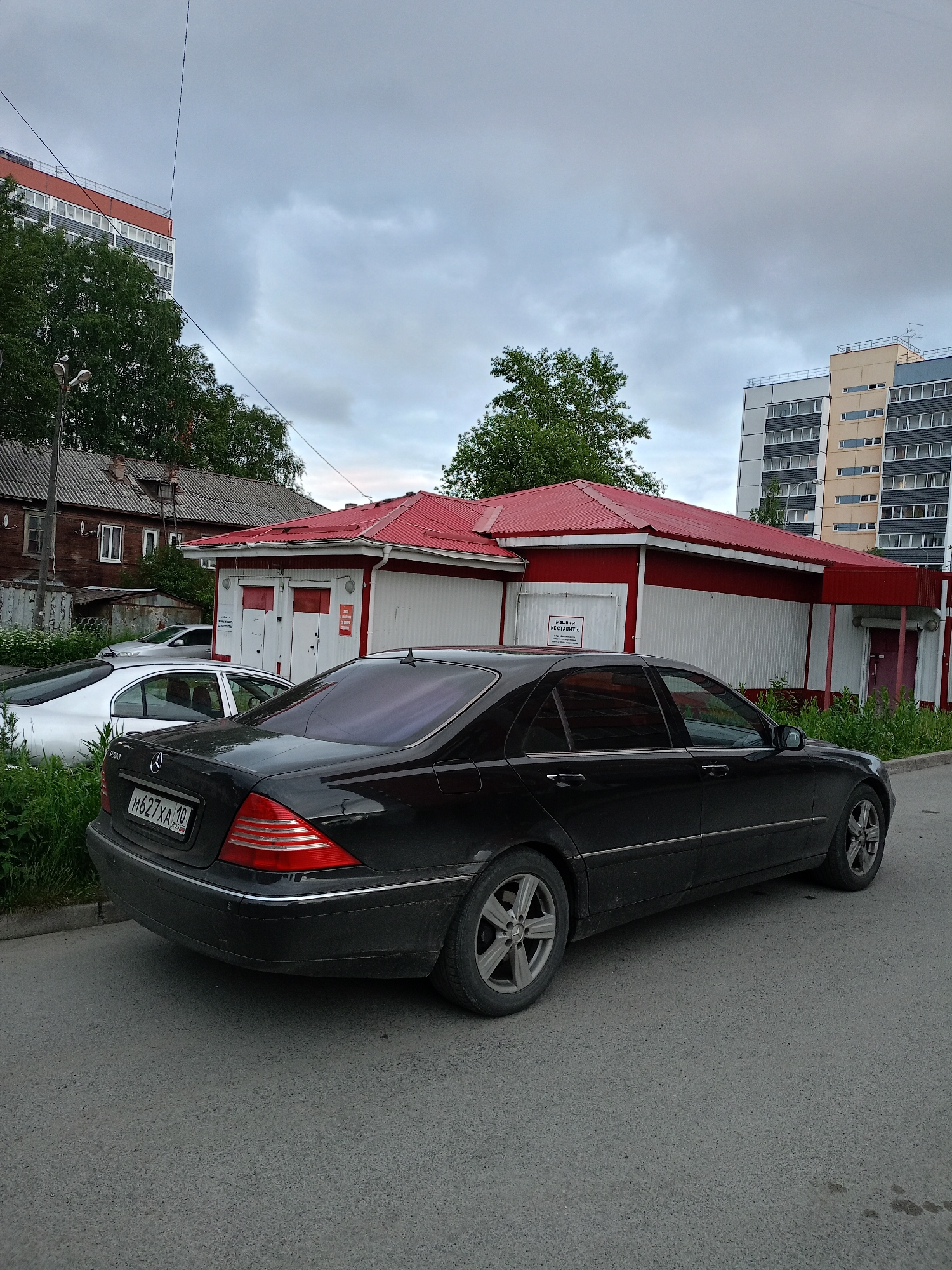 The image shows a black Mercedes-Benz S-Class sedan parked on a paved street in front of a building with a red roof. The building appears to be a small commercial structure, possibly a kiosk or a small shop, with white and red siding. There is a sign on the building that reads "МАШИНЫ НЕ СТАВИТЬ!" which translates to "DO NOT PARK CARS!". In the background, there is an older wooden house and a modern apartment building. The sky is overcast. The license plate on the Mercedes-Benz reads "M627XA 10 RUS". The image was taken in Petrozavodsk, Russia.