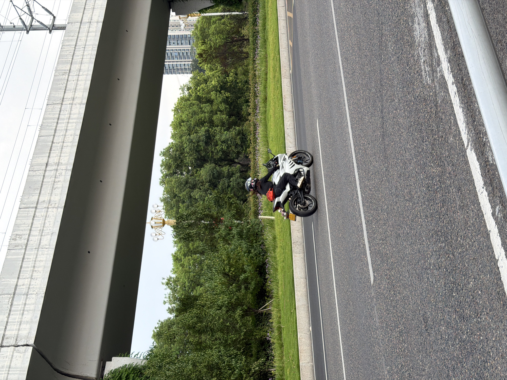 A motorcycle rider wearing a helmet and a black top is on a white motorcycle, traveling on a paved road. The rider is carrying a red bag on their back. The road has white lane markings. In the background, there is a row of green trees, a grassy area, and a section of a concrete overpass. Beyond the trees, a tall building is visible. The sky is overcast. The scene appears to be in Jiangjin, China, indicated by the presence of typical urban infrastructure like overpasses and modern buildings, alongside lush greenery.