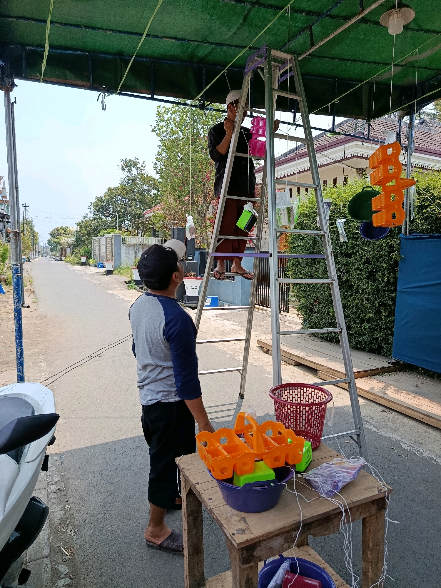 In Bululawang, Indonesia, two men are setting up decorations under a green awning. The man on the left, wearing a black long-sleeved shirt, white cap, and sarong, is on a silver A-frame ladder, attaching pink plastic decorative elements to strings hanging from the awning. The man on the right, wearing a grey and blue long-sleeved shirt, black cap, and black pants, stands on the ground, looking up at the man on the ladder.
In the foreground, a wooden table holds a purple bowl containing green and orange plastic items, and a plastic bag of what appear to be small, colorful objects. A red plastic basket is also present on the ground near the table. To the left of the table, the back of a white motorcycle is visible. The background shows a residential street with houses and lush green foliage. The overall scene suggests preparation for an event or sale.