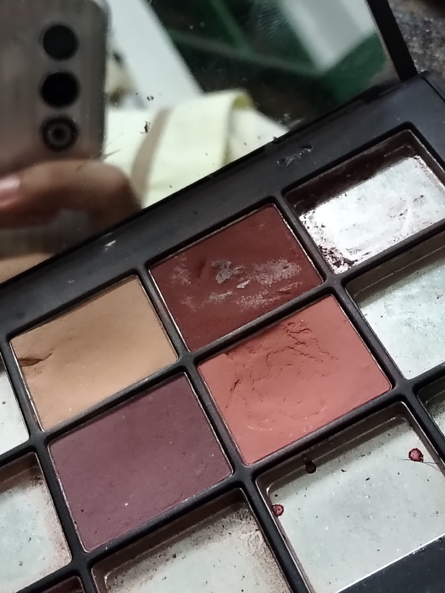 The image shows a close-up of an eyeshadow palette. The palette is black and contains several rectangular pans of eyeshadow. Some of the pans are full, while others are partially used. The eyeshadow colors visible include a light brown, a reddish-brown, a lighter reddish-orange, and a deep burgundy. There are also some pans that appear to be lighter, possibly with a shimmer or metallic finish, though they are not clearly defined. The surface of the eyeshadows shows signs of use, with some powderiness and indentations.
In the reflection on the lid or a mirror above the palette, a person's hand and a smartphone with three circular lenses are visible. The setting appears to be indoors, with a slightly out-of-focus background that suggests a casual environment. There is no visible text in the image. The lighting suggests it could be daytime, but it's difficult to determine the exact time or weather conditions. The overall impression is of a personal makeup item that has been used.