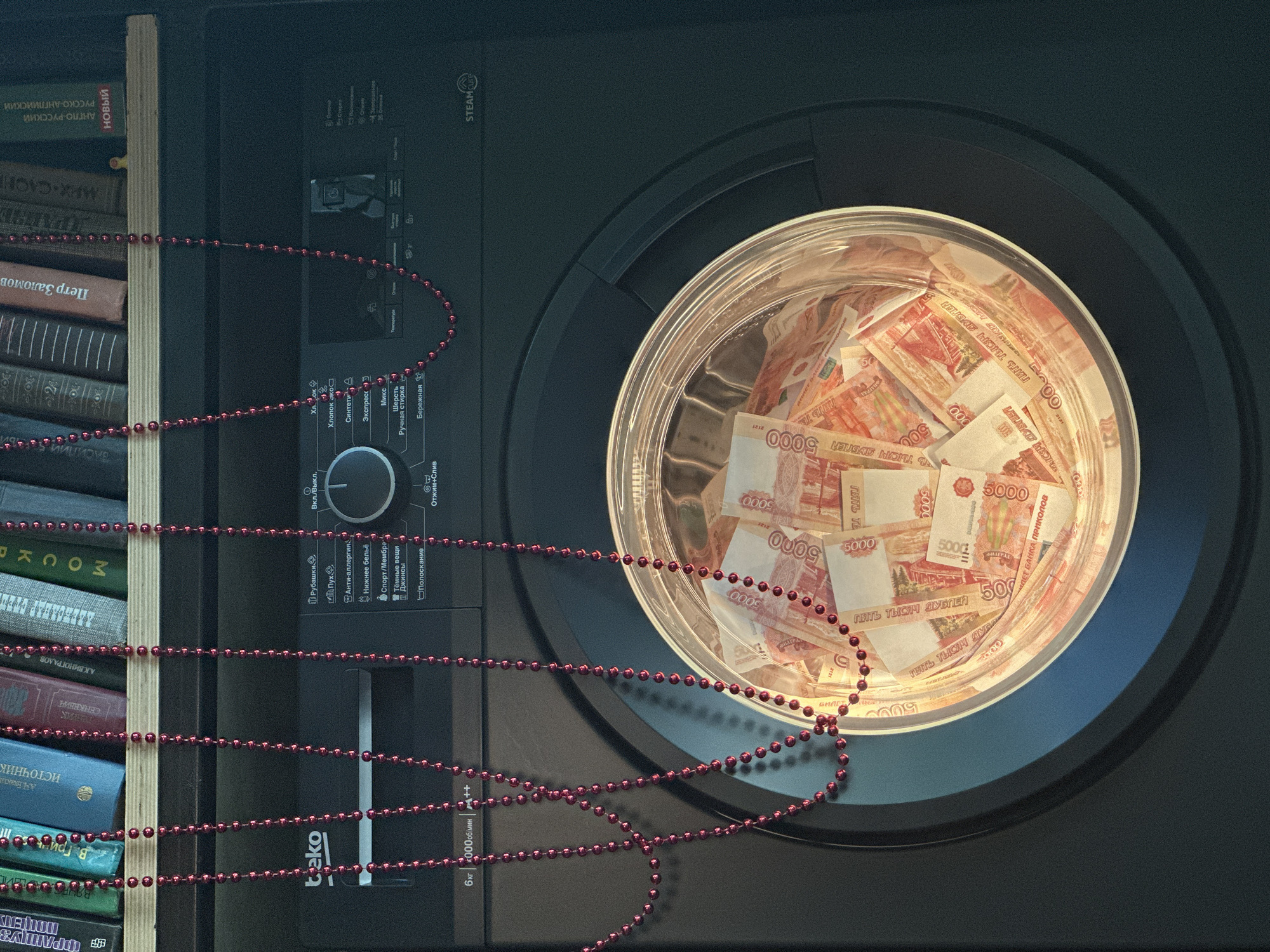 The image shows a washing machine containing a pile of Russian 5000-ruble banknotes. To the left of the washing machine is a stack of books on a shelf. Red bead necklaces drape across the books and the front of the washing machine. The washing machine is labeled "beko" and has various washing cycle options printed in Russian. Some of the book titles are also in Russian. The location in the city of Novosibirsk, Russia, cannot be determined from the given image.