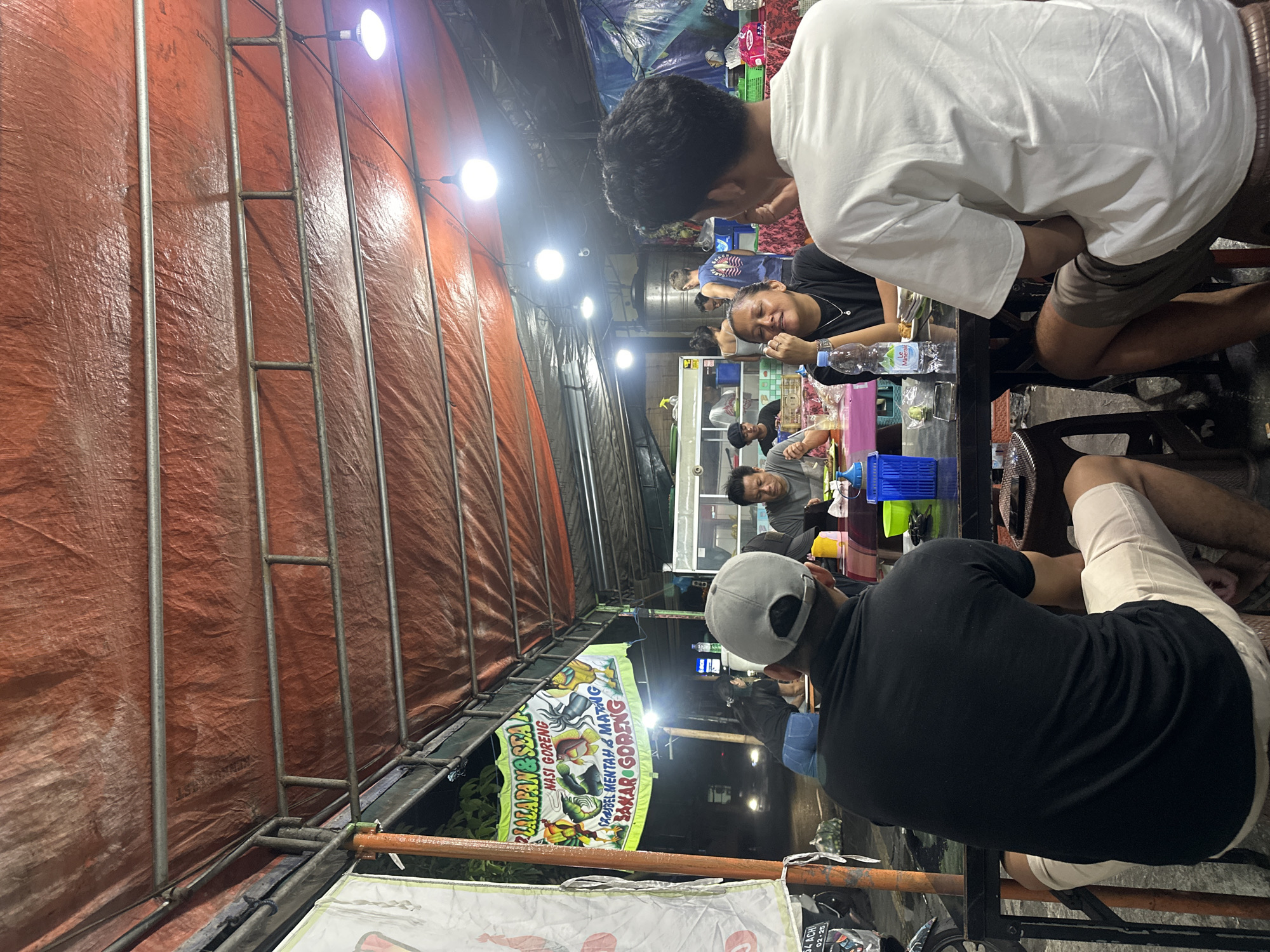 The image depicts an outdoor eating area in Denpasar, Indonesia. Diners are seated at a black table. The foreground shows the back of a man in a black shirt and tan shorts wearing a grey baseball cap. Other people are seated at the table, including a woman in a black shirt and a man in a white shirt. A partially visible advertisement banner displays the words "LALAPAN&SEA NASI GORENG". An orange tarp with a metal frame forms the overhead and side covering of the area. The surroundings include a city street at night.