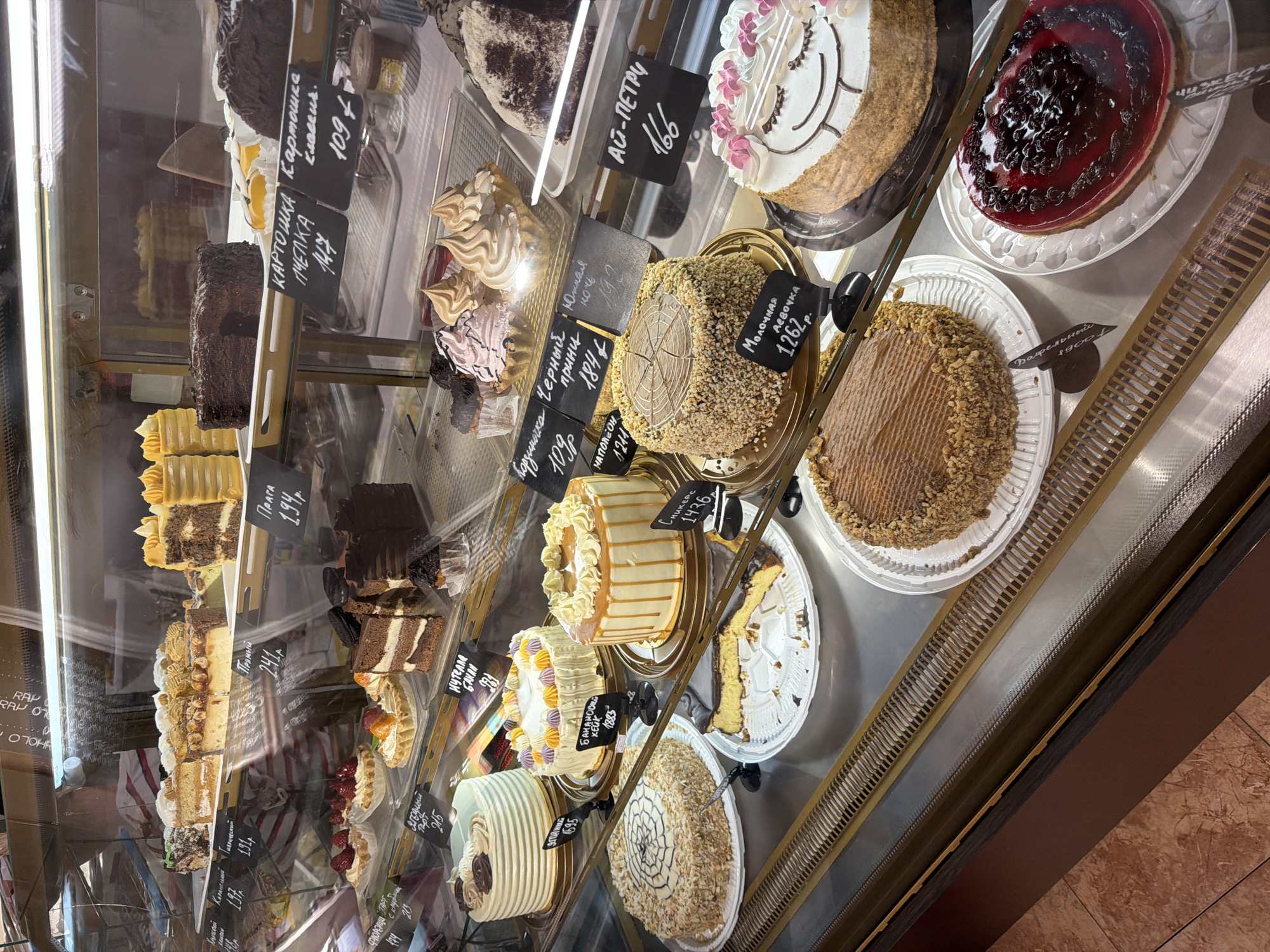 The image shows a refrigerated display case in a bakery, filled with a variety of cakes and pastries. The bakery appears to be located in Alushta, Ukraine, as indicated by a price tag with Cyrillic script and the currency symbol "₽" which is commonly used in Russia and some other countries, but given the specified location, it's reasonable to assume it might be local currency or a common display practice.
In the top shelf, several cakes are visible, including what appear to be layered cakes with chocolate and caramel frosting, and some smaller, individual pastries. Below this, there are more cakes, including tarts topped with strawberries and fruit, and chocolate cakes.
The middle shelves showcase larger, decorated cakes. One cake is white with intricate frosting patterns, another is yellow with caramel drizzles and piped frosting, and a third is round and covered in nuts with a wood-grain pattern on top. There is also a cheesecake with a chocolate topping on a white plate, and a slice already removed.
The bottom shelf features more cakes, including a round cake covered in nuts and a heart-shaped cake with a dark red glaze and berries on top.
Several small, black price tags with handwritten text in Cyrillic are placed in front of the baked goods, indicating prices and names of the items. The environment is clean and well-lit, typical of a retail food establishment. No people are visible in the frame.