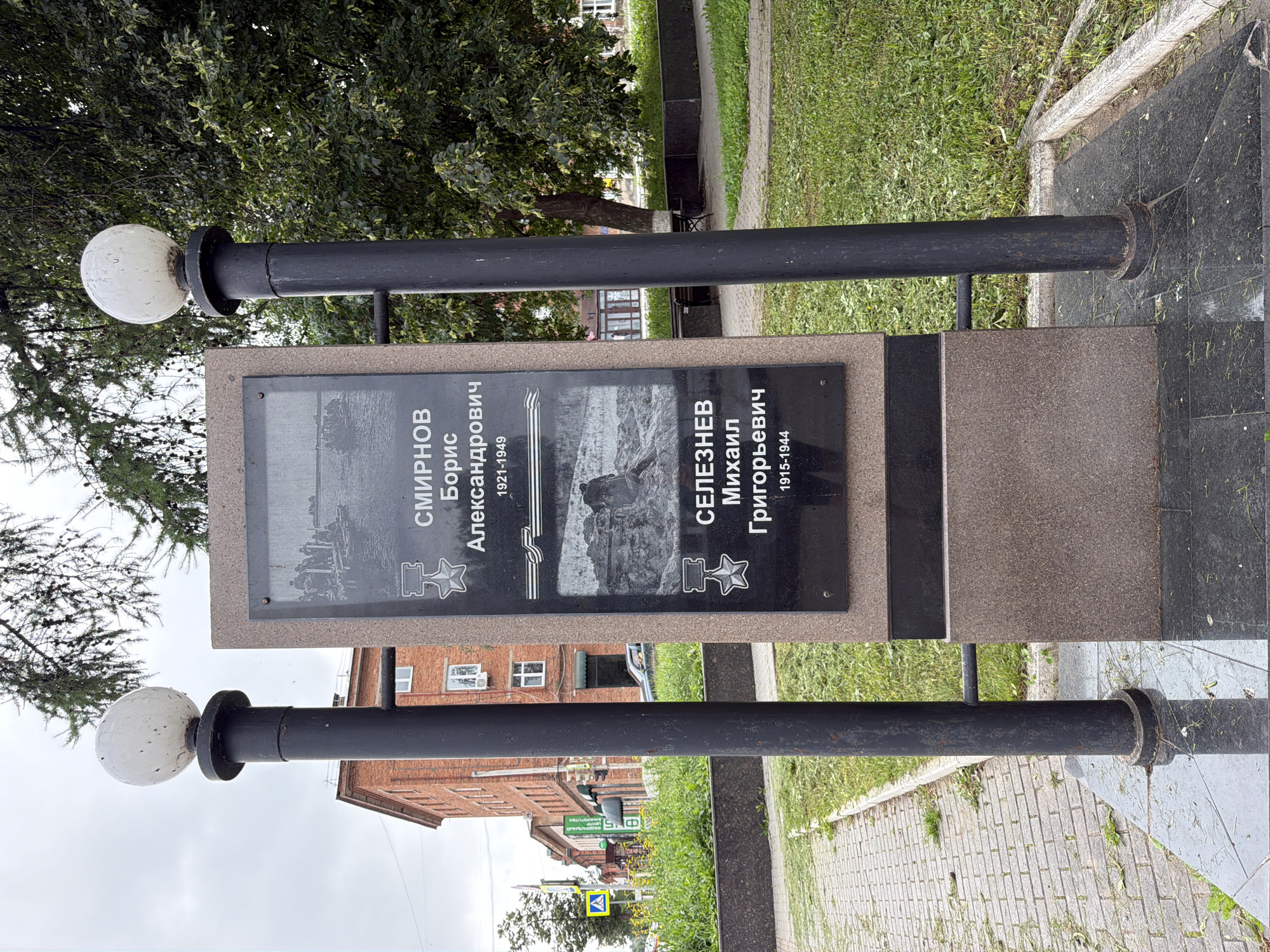 The image shows a war memorial in Votkinsk, Russia. The memorial consists of a granite plaque with the names and life dates of two individuals, SMIRNOV Boris Alexandrovich (1921-1949) and SELEZNEV Mikhail Grigorievich (1915-1944), along with their portraits and military decorations. The memorial is flanked by two black lampposts topped with white globes. The surrounding environment includes a paved walkway, green grass, trees, and buildings in the background. The sky is overcast.