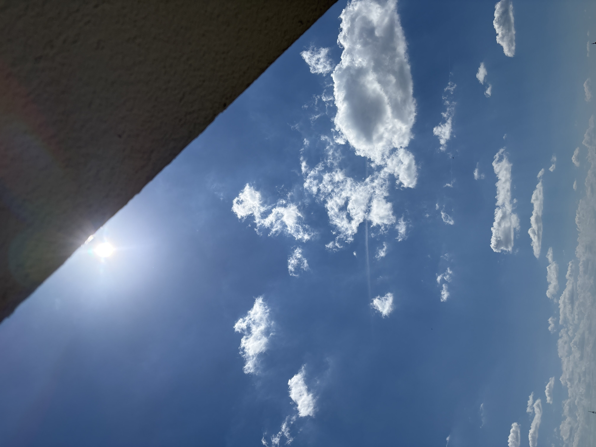 The image captures a bright, sunny sky with scattered white cumulus clouds over Bönnigheim, Germany. The sun is high and prominent in the upper left quadrant, casting a glare. A textured, light-colored architectural element, possibly a building overhang or a wall, occupies the upper right portion of the frame, diagonally bisecting the view. The lower half of the image is dominated by the clear blue sky and the various cloud formations, some of which appear fluffy and well-defined, while others are wispy. There are no people or visible subjects in the frame, only the sky and the suggestive edge of a structure. The scene depicts a typical daytime sky, possibly from an outdoor vantage point within the city.