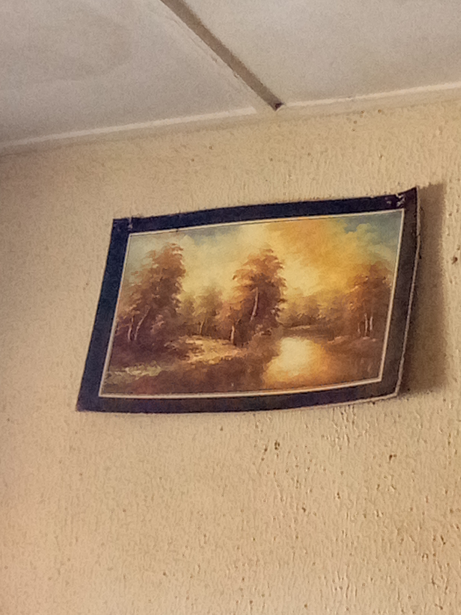 A framed landscape painting of trees and a body of water hangs on a textured, light-colored wall. The painting depicts a serene scene with warm, diffused lighting, suggesting either sunrise or sunset. The frame appears to be dark blue or black. The wall itself has a rough, slightly bumpy texture, and a horizontal line is visible across the top of the image, likely indicating a ceiling or architectural detail. The lighting in the scene is artificial, casting a slight shadow on the painting from an unseen light source. The painting is angled slightly, with the top edge dipping lower than the bottom edge. The overall impression is of a domestic indoor setting. The location is not specified in the image.