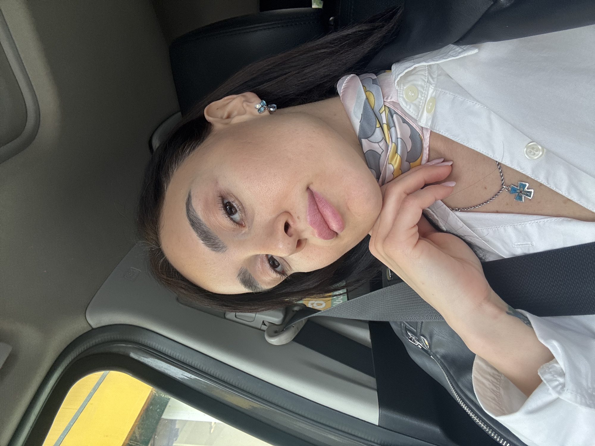A woman is in a car, wearing a white shirt, a black jacket, and a necklace with a blue cross. She is also wearing an earring with blue stones. She has dark hair and eyebrows. She is sticking her tongue out. The car is moving, as indicated by the blurred yellow building seen through the window. The exact location in Tbilisi, Georgia, cannot be determined from the image alone.