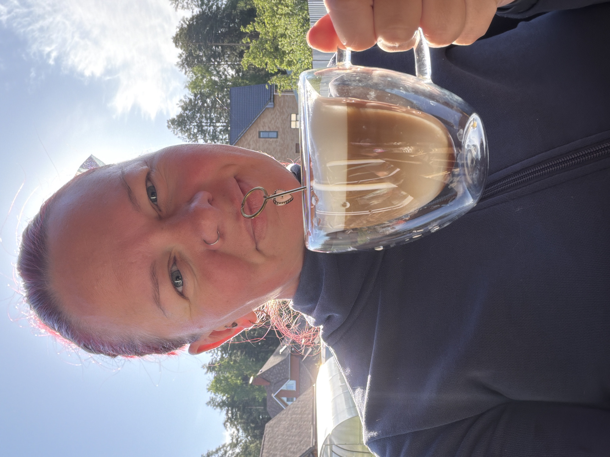 A selfie taken outdoors. A person with pink and purple hair holds a clear, double-walled glass mug filled with a beverage that appears to be a latte, with distinct layers of white and brown liquid. The person has multiple facial piercings, including a nose ring and a lip piercing that appears to have a small, ornate adornment. They are wearing a dark blue zippered jacket or sweatshirt. In the background, blurred details suggest a suburban or rural setting. There are houses with varying roof styles and greenery including trees visible. Considering the request for the location to be Chaikovsky, Russia, these structures and the overall setting align with typical suburban environments in that region. The sky is visible and sunny.