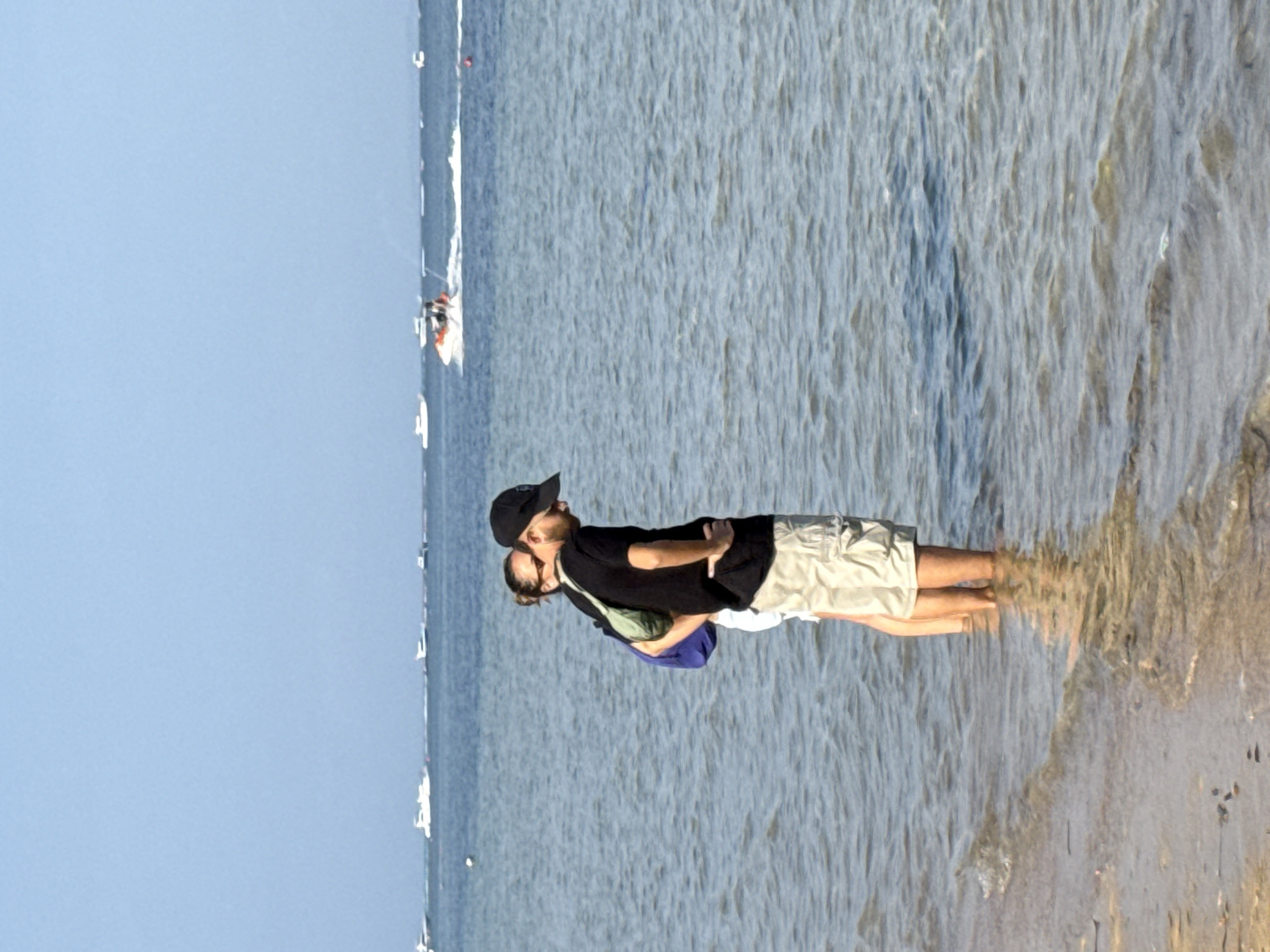 A man and a woman stand in shallow water on a beach, with their feet submerged. The man, wearing a black t-shirt and beige shorts, stands facing away from the camera with his head down. The woman, wearing a blue top and white shorts, stands behind and to the left of the man, looking towards him. The water is calm with gentle waves lapping at the shore. In the background, several boats are visible on the horizon, including a jet ski in motion. The sky is clear and blue. The scene suggests a relaxed day at a beach location, possibly in Denpasar, Indonesia, known for its coastal areas and water activities.