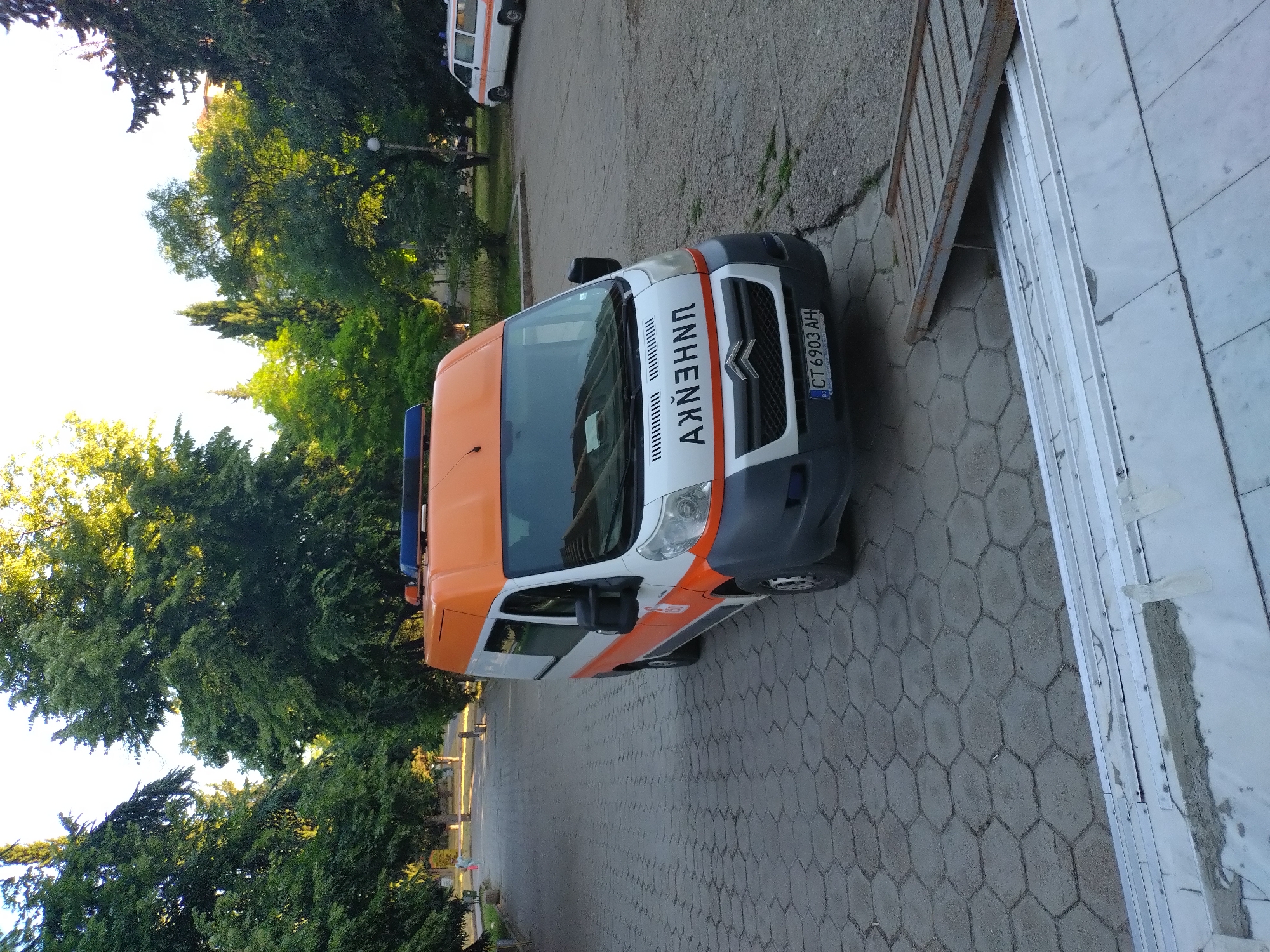 The image shows an ambulance van parked on a hexagonal-patterned paved walkway in Stara Zagora, Bulgaria. The van is white and orange, with the word "АНBULANЦ" (ambulance) written in black Cyrillic letters on the front. The license plate reads "CT 6903 AH". Trees line the walkway, and another white van is visible in the background. The scene appears to be a public area, possibly a park or hospital grounds, in the late afternoon or early evening, given the lighting. A ramp is positioned next to the ambulance.