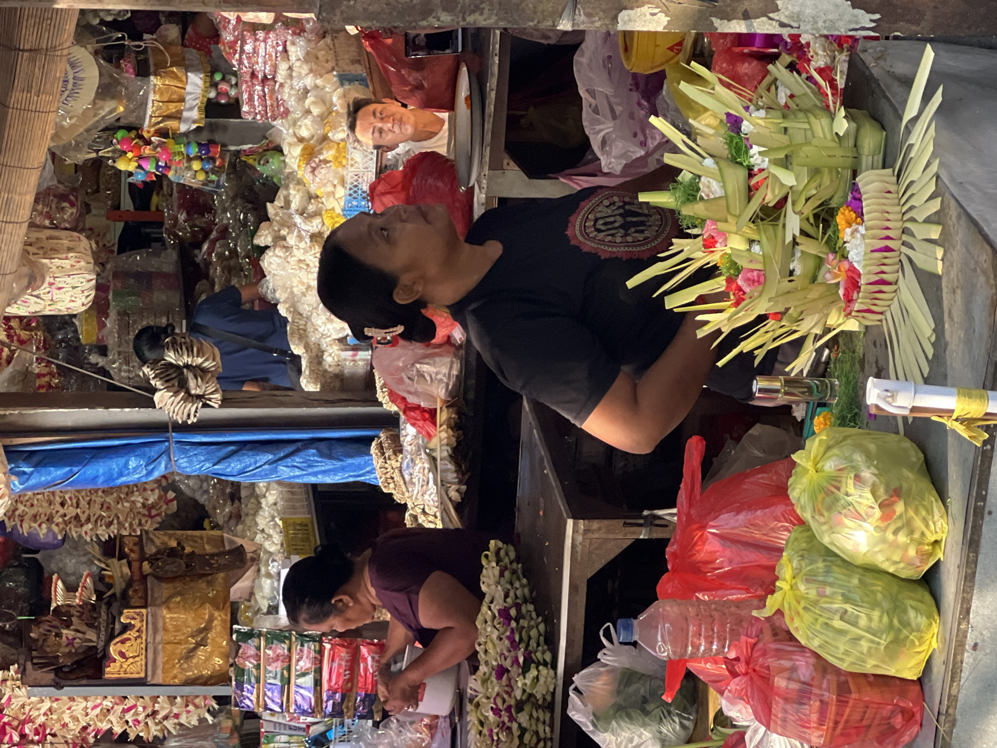 A medium shot captures an outdoor market scene in Ubud District, Indonesia, with a focus on two women preparing and displaying floral offerings. In the foreground, a woman in a black t-shirt with a circular logo on the chest is actively arranging intricately folded green leaf baskets filled with colorful flowers. To her left, another woman in a maroon shirt is working with a white bowl and appears to be sorting or preparing flowers. The background is filled with various market stalls, displaying an abundance of goods, including stacks of packaged snacks, woven baskets, and floral decorations hanging from above. Several large, translucent plastic bags containing flowers and greenery are also visible, some red and some yellow-green. A man's face is partially visible in the mid-ground, looking towards the right. The overall atmosphere is that of a busy, traditional market setting.