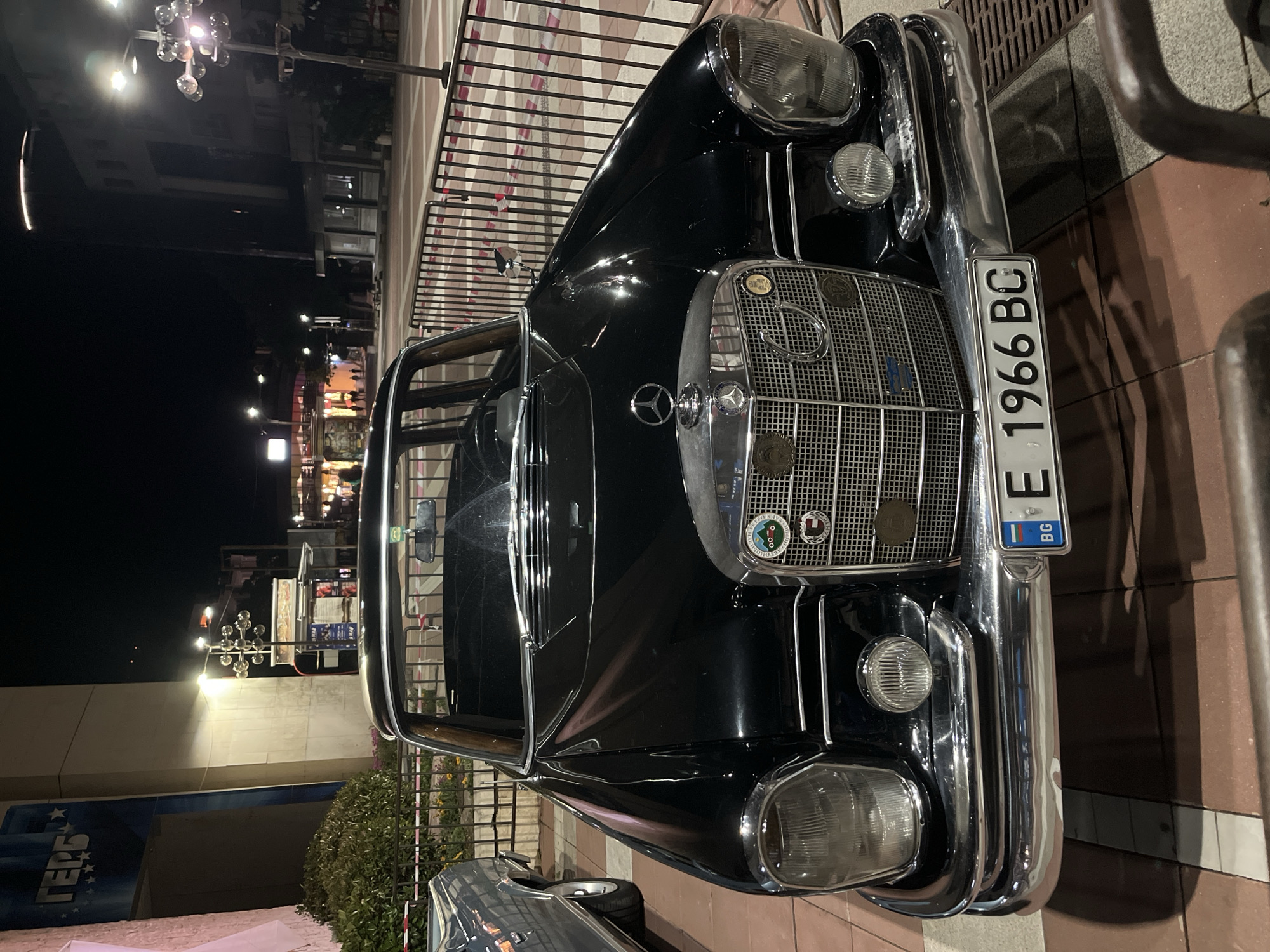 The image depicts the front of a classic black Mercedes-Benz car parked on a patterned pavement at night. The car features a prominent grille with the Mercedes-Benz logo and decorative badges, round headlights, and chrome bumpers. A Bulgarian license plate with "E 1966 BC" is visible on the front bumper. The background shows a street scene with buildings, streetlights, and a metal railing. The location appears to be a well-lit urban area, but the specific city and location are not discernible from the image.