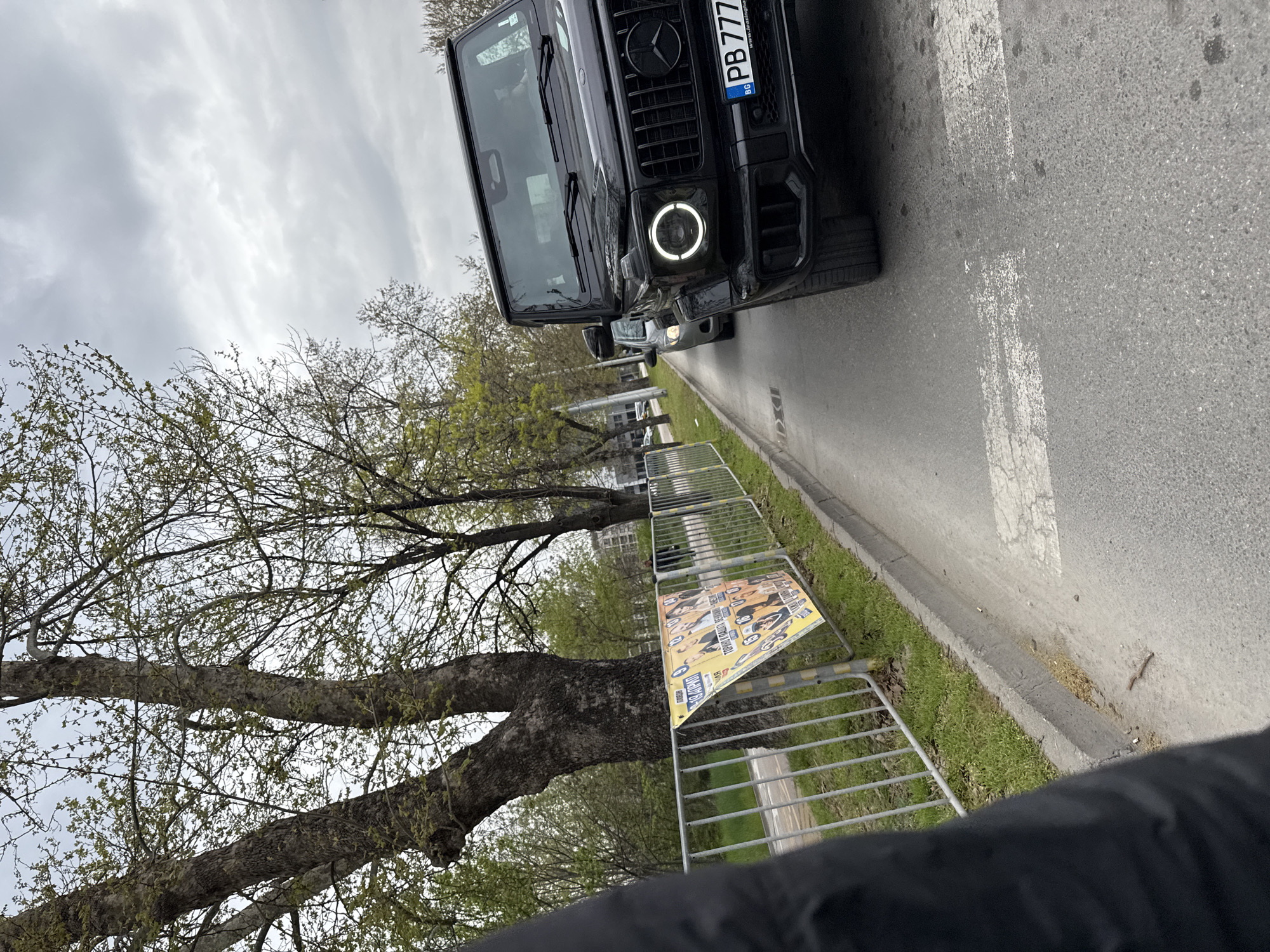 The image shows a black Mercedes-Benz G-Class parked on the side of a road in Plovdiv, Bulgaria. The car's front is visible, and its license plate reads "PB 7777 BT". To the left of the vehicle, there is a tree with budding green leaves and a metal crowd control barrier. A poster is attached to the barrier. The sky is overcast. The location is along a road with a sidewalk and green space.