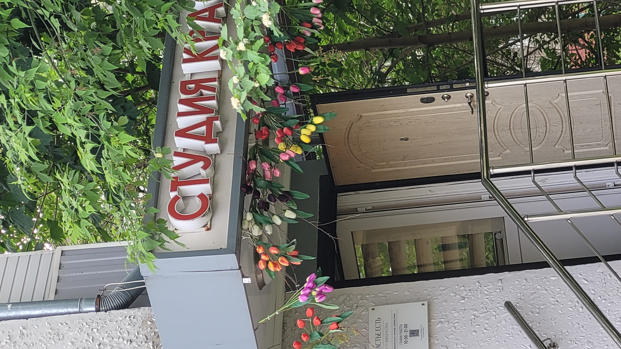 Here is a description of the image:
The image shows the exterior of a building in a city labeled as Unknown, Unknown. A vertical sign with the word "СТУДИЯ" in red letters is prominently displayed on the building's facade. Decorative elements, including artificial flowers in various colors (red, pink, yellow, purple, orange, and white tulips), adorn the building's exterior, seemingly wrapped around the sign and parts of the building. A door with decorative patterns is visible above the sign. A stainless steel railing extends along the side of the building. A sign with written information is attached to the building, below the decorative flowers. Green foliage overhangs parts of the building.