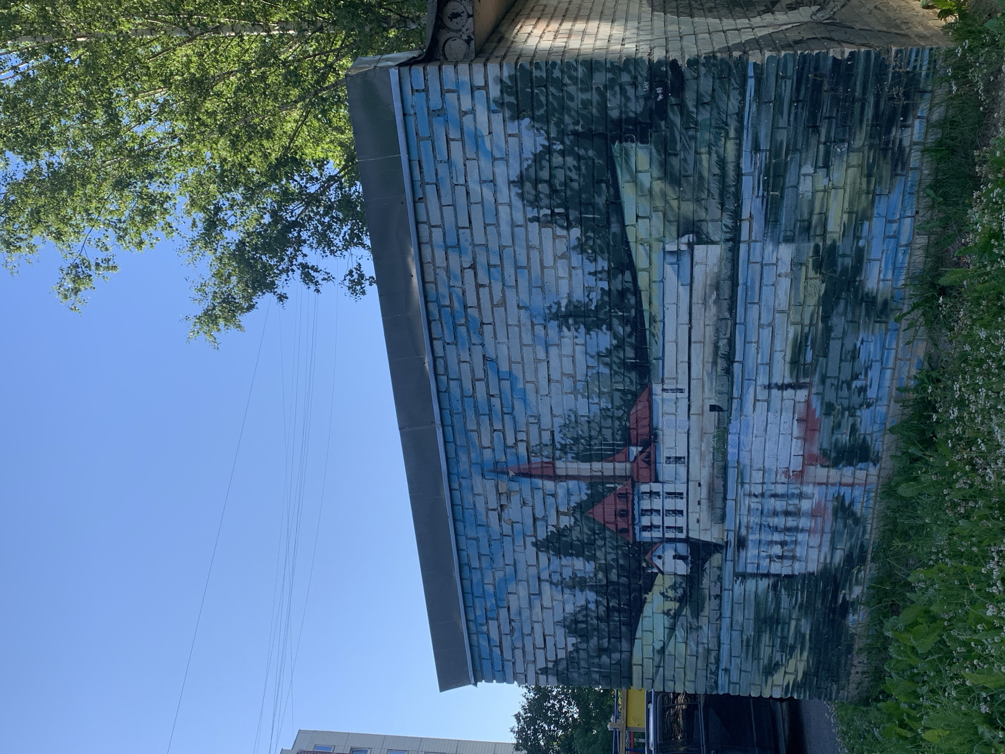The image shows a mural painted on the side of a brick building. The mural depicts a landscape with a body of water in the foreground and a building with red roofs and a tall red spire in the background, surrounded by trees. The reflection of the building and trees is visible in the water. The building itself appears to be a shed or utility structure, with a metal roof overhang. The brickwork is visible beneath the paint, indicating it is a mural on a real wall. The surrounding environment is green and overgrown with grass and small white flowers. In the background, a tall apartment building and leafy trees are visible against a clear blue sky with some power lines. This mural is located on the side of a building in the park area of Gatchina, Russia, likely near the Gatchina Palace.
