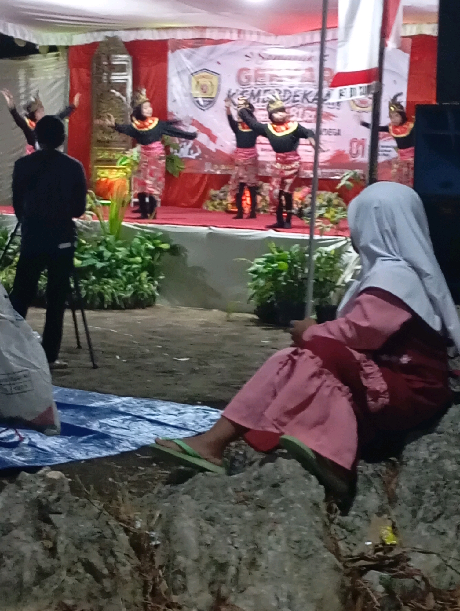 In the city of Ketepung, Indonesia, a group of young girls in traditional Indonesian attire perform a dance on a stage draped with red and white banners. The banners display text, some of which is visible, including "GEMPITA KEMERDEKAAN" and the number "01". A man stands with his back to the camera, observing the performance. In the foreground, a woman wearing a light-colored headscarf and a pink dress sits on a rocky area, facing away from the stage. The ground is covered with dirt and scattered debris. Lush green foliage is visible on the left side of the stage.